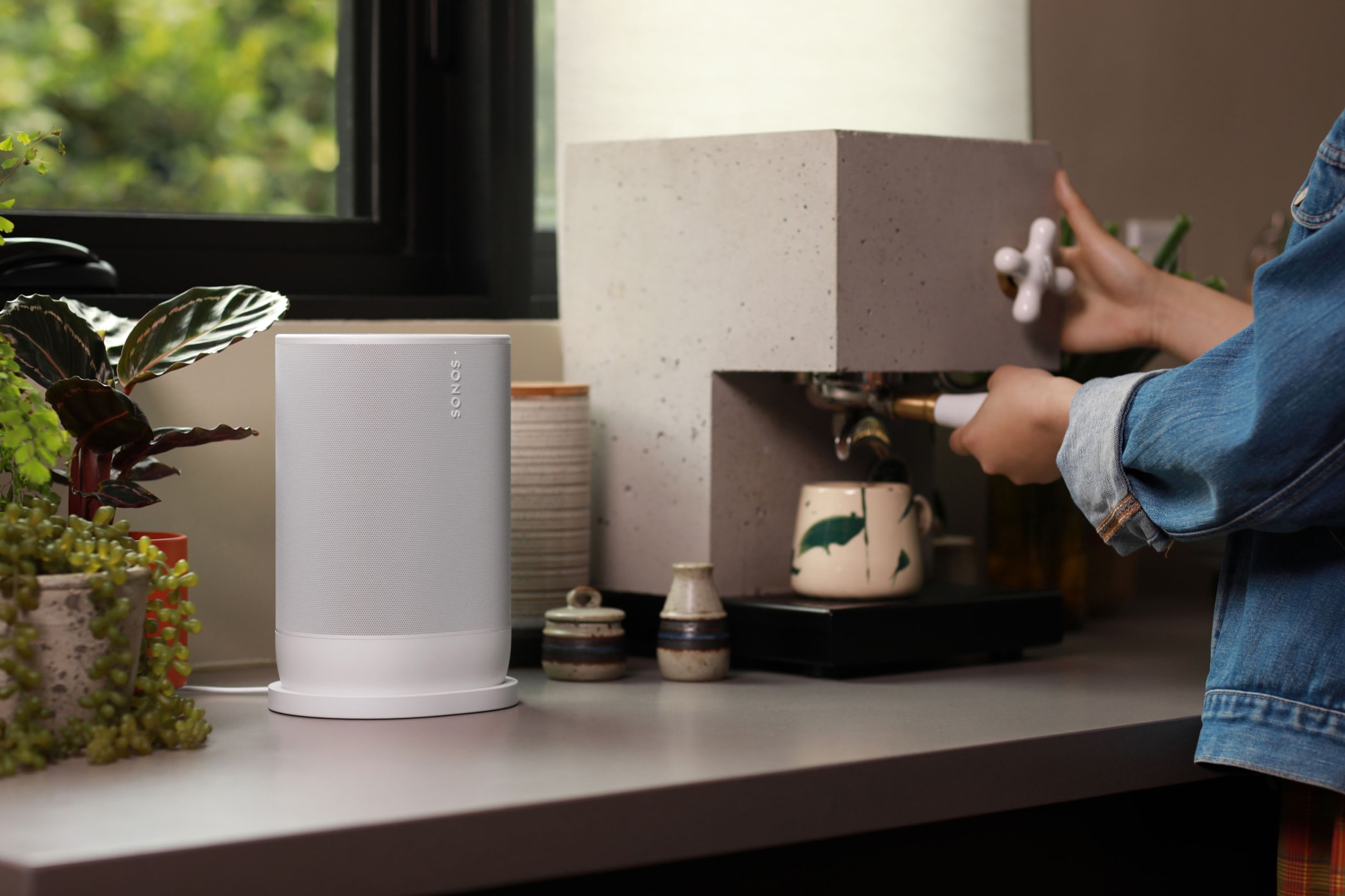 A photo of a woman making espresso at the kitchen counter next to a white Sonos Move 2