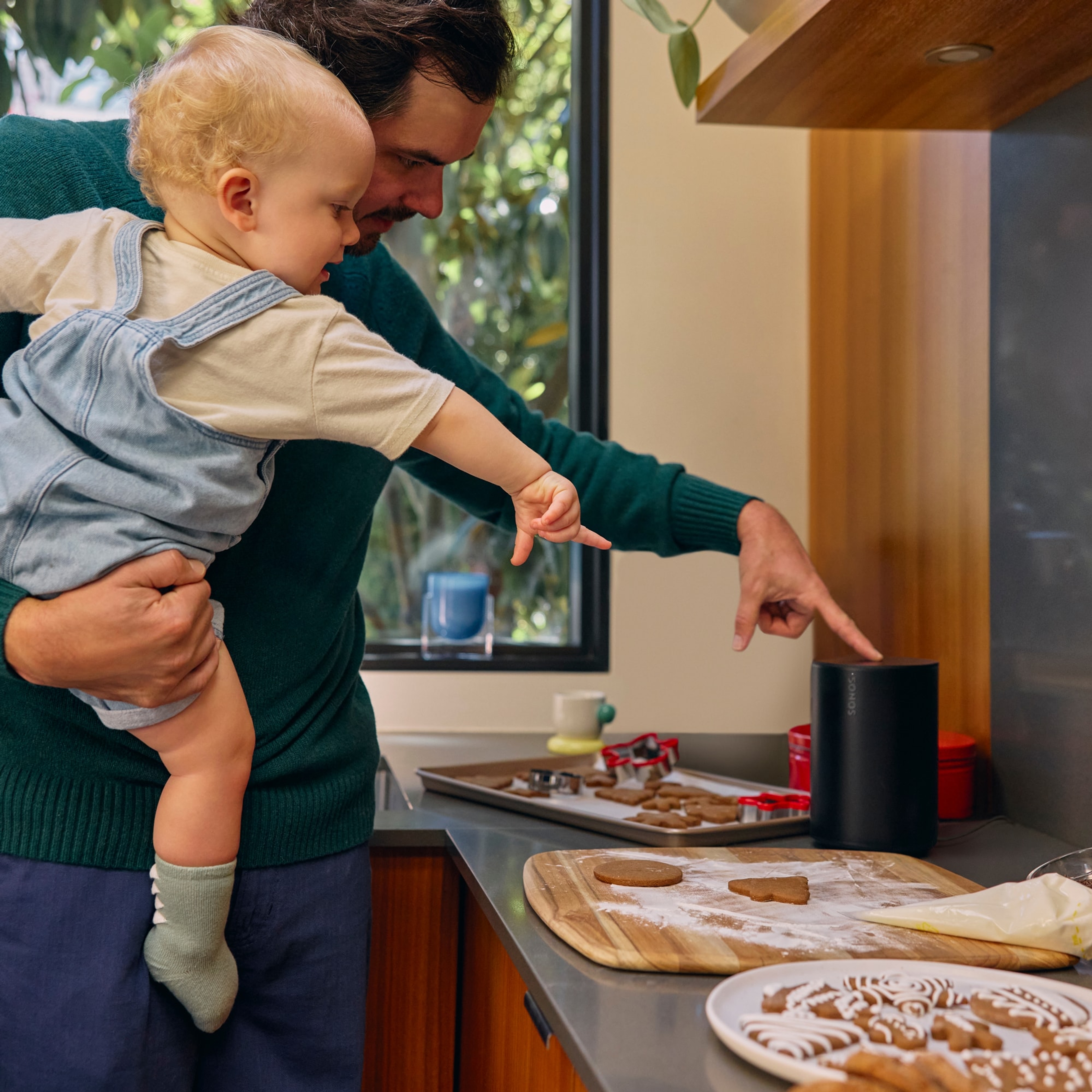 A dad holding his infant while making holiday cookies listening to a black Sonos Era 100 speaker