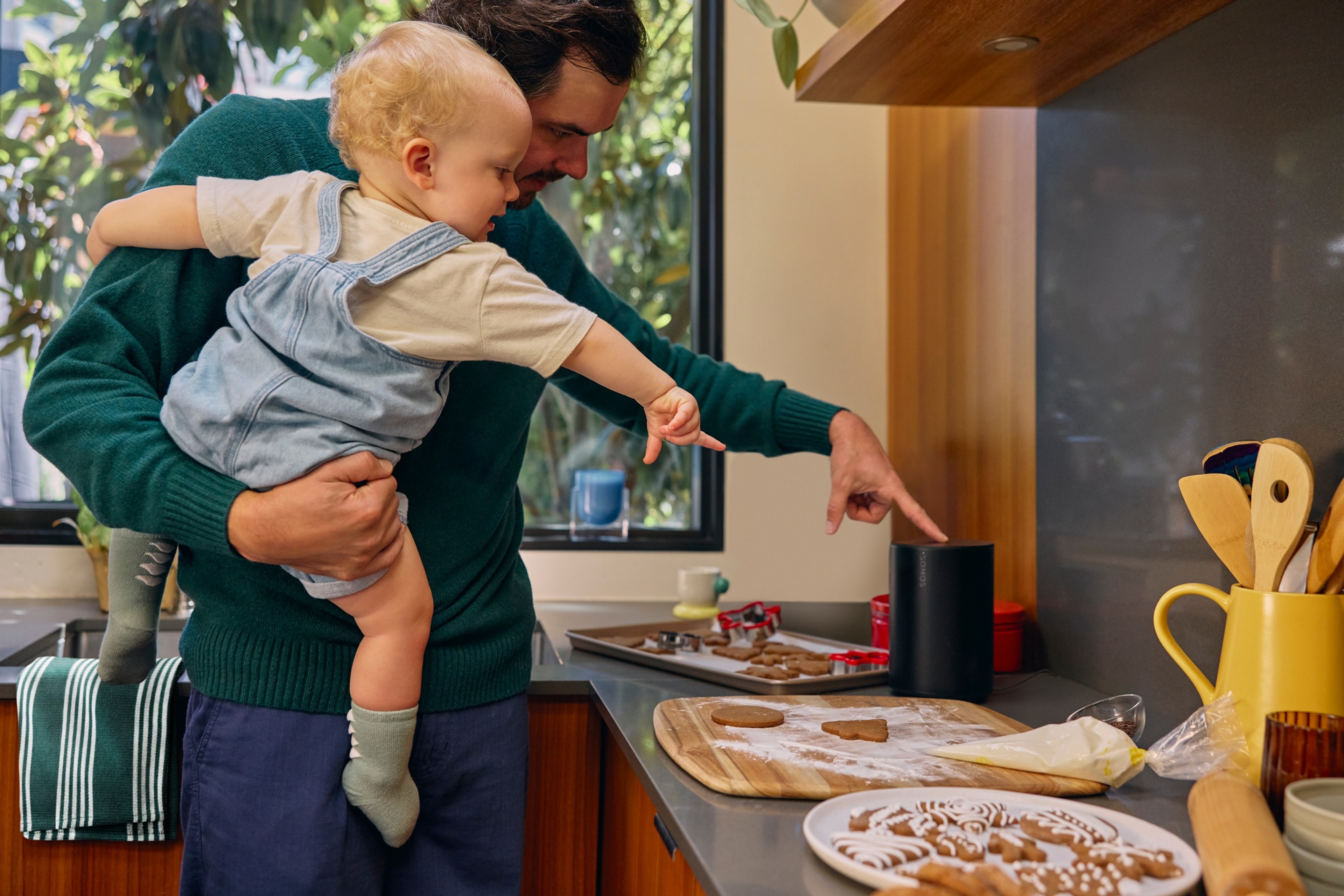 A dad holding his infant while making holiday cookies listening to a black Sonos Era 100 speaker