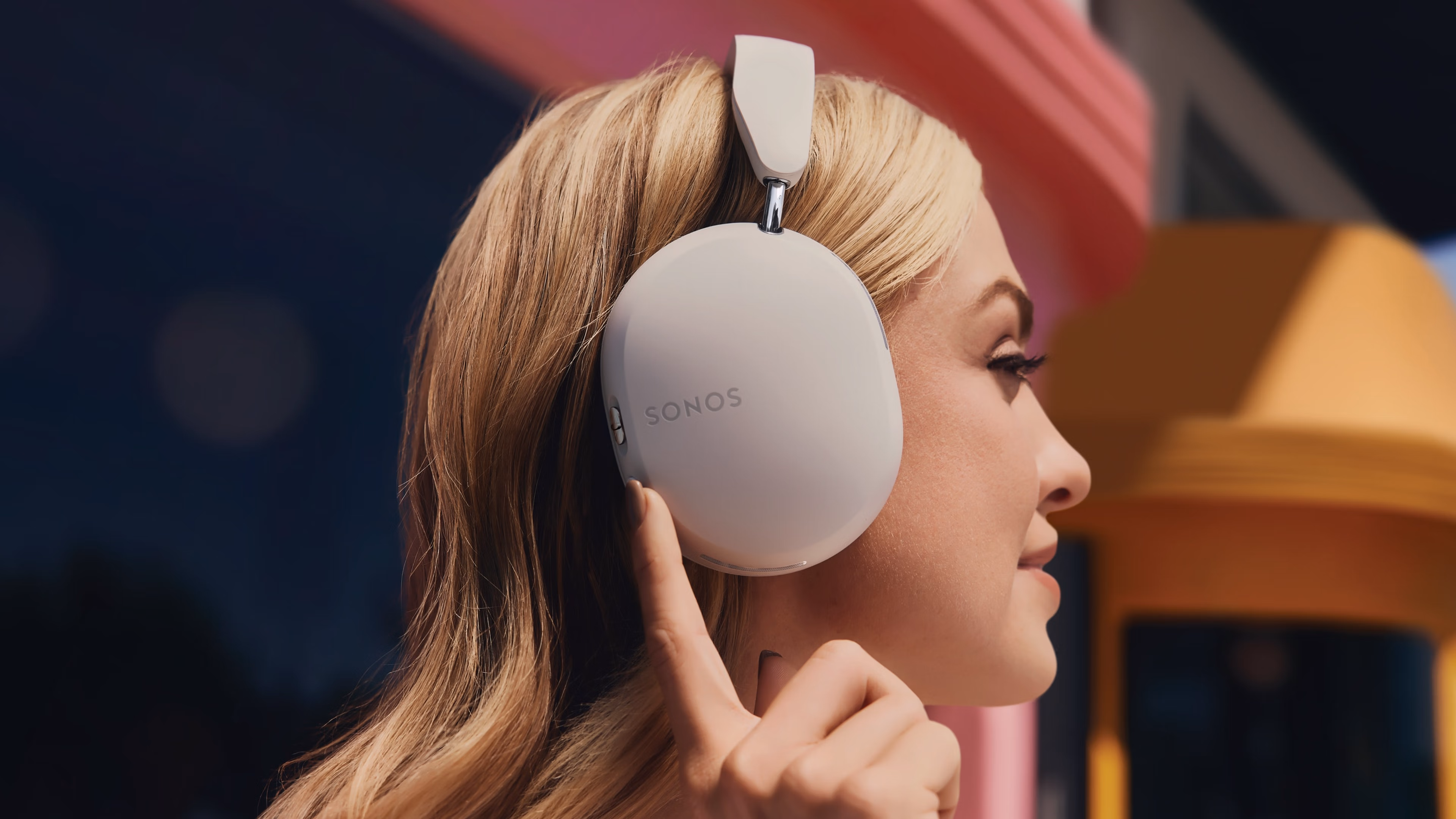 Side profile of a person wearing Soft White Sonos Ace headphones and pressing the active noise cancellation button
