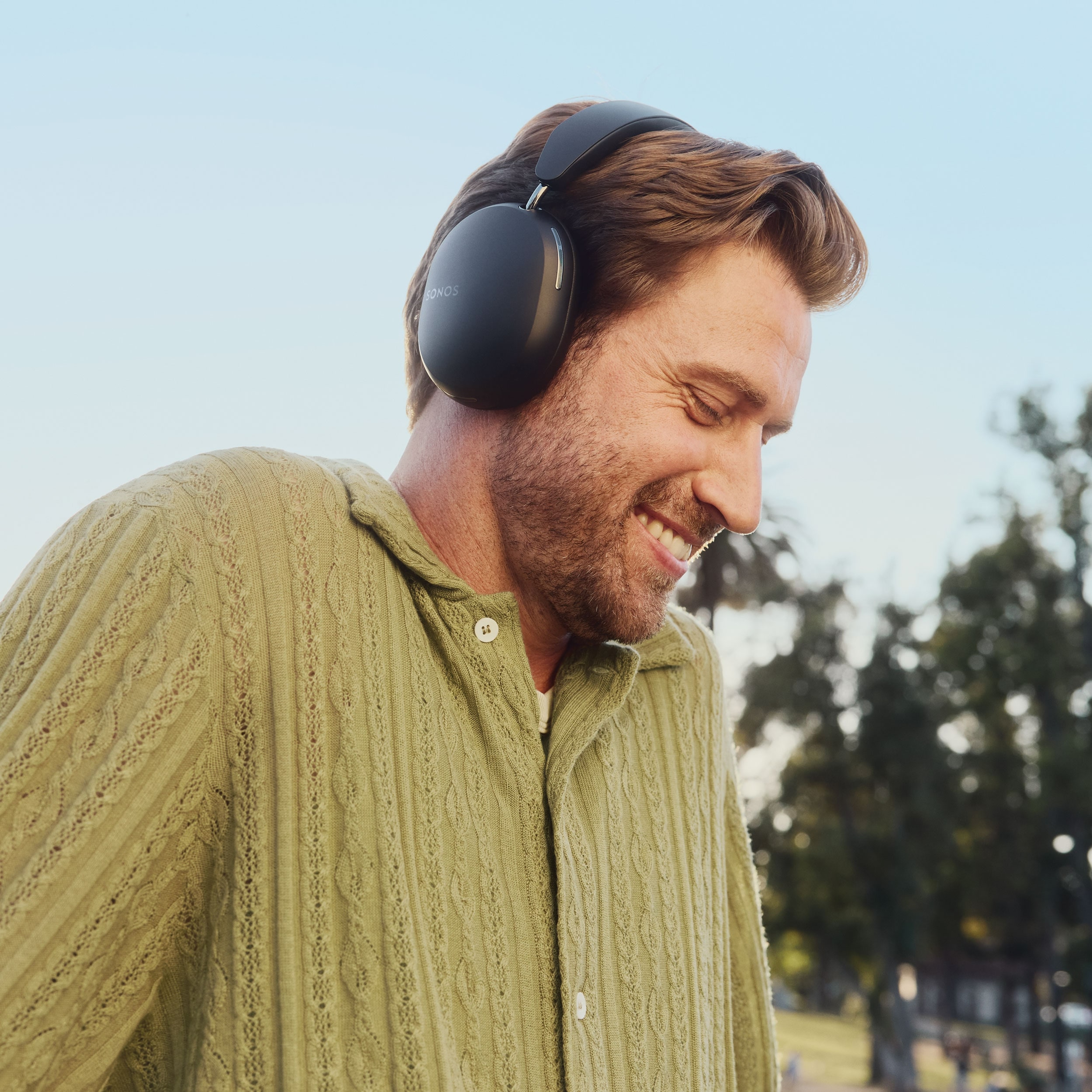 Una persona sonríe y escucha música en sus audífonos Sonos Ace en negro mientras camina por el parque