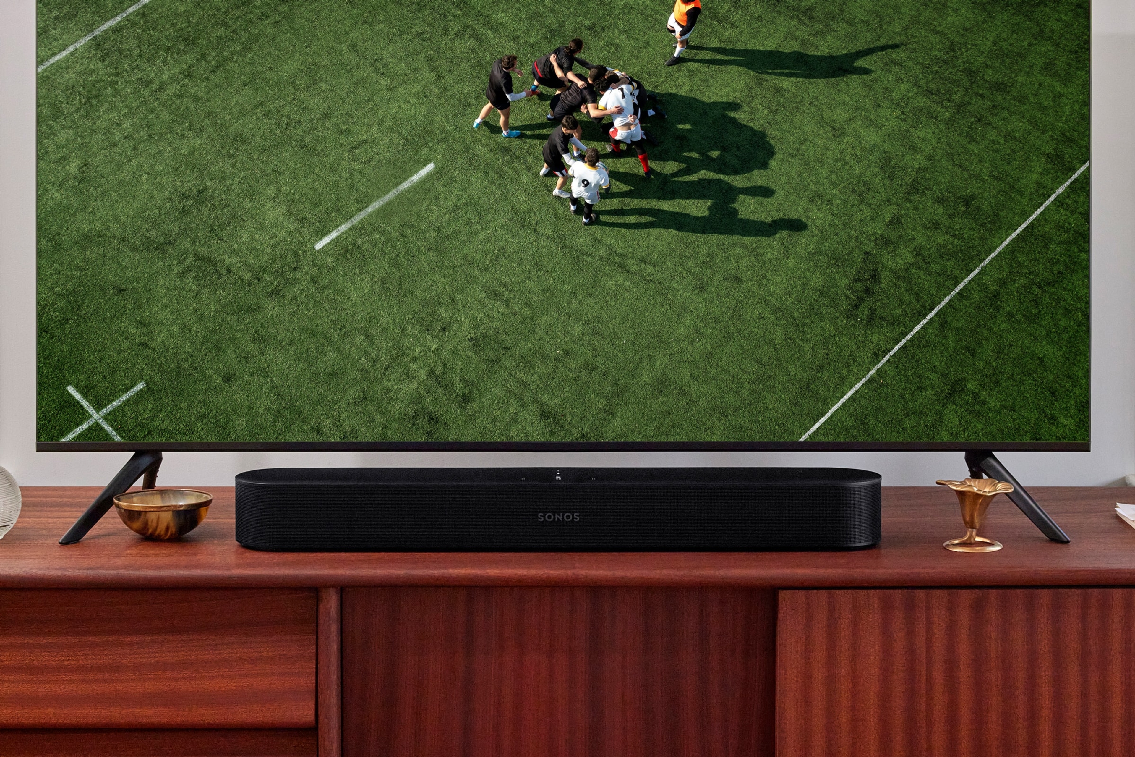 Black Sonos Beam on a credenza below a TV showing a Rugby match