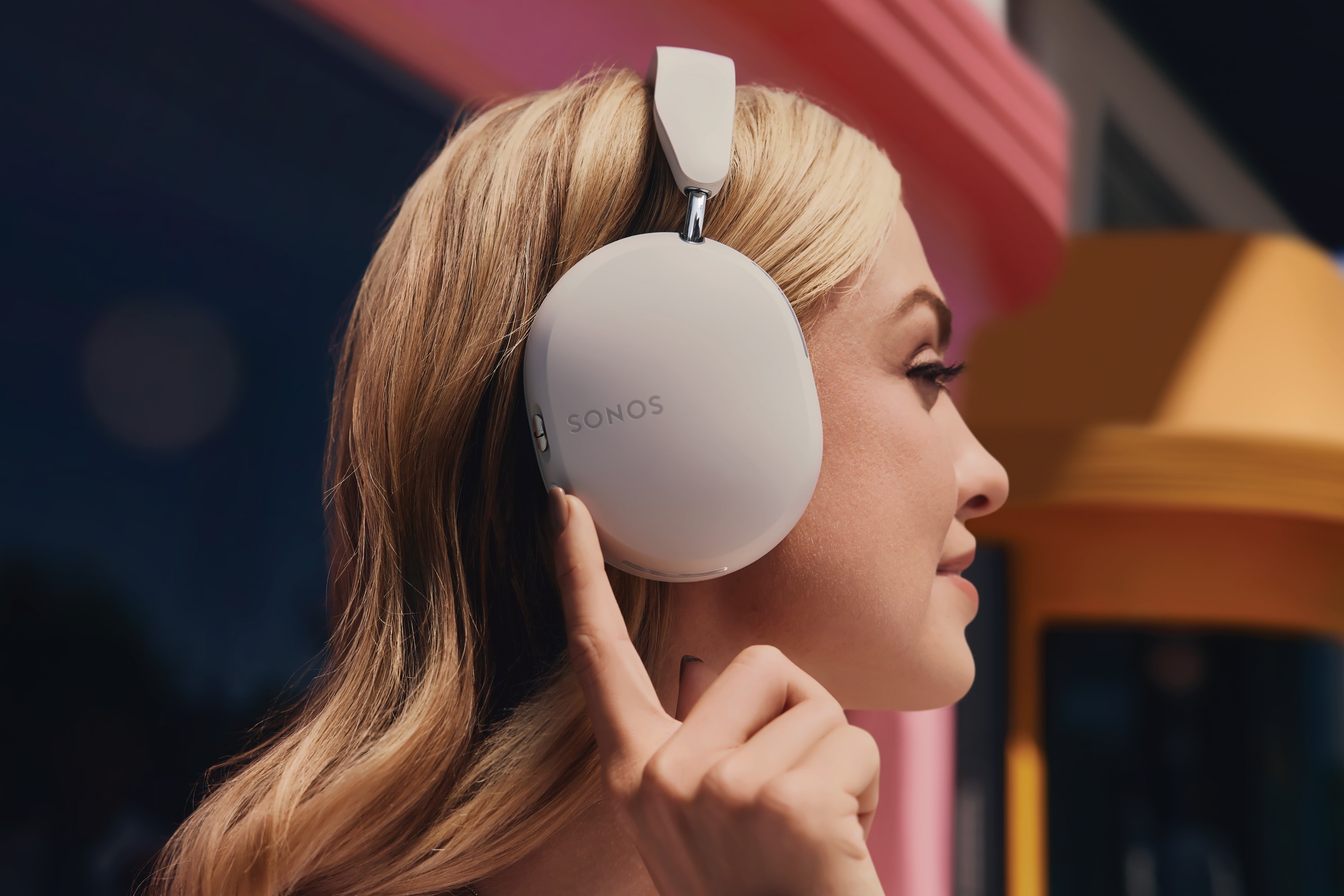 Side profile of a person wearing Soft White Sonos Ace headphones and pressing the active noise cancellation button