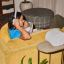 Person sitting on a yellow couch listening to music on white Sonos Ace Ultra headphones next to a table with a white Era 300 speaker on it