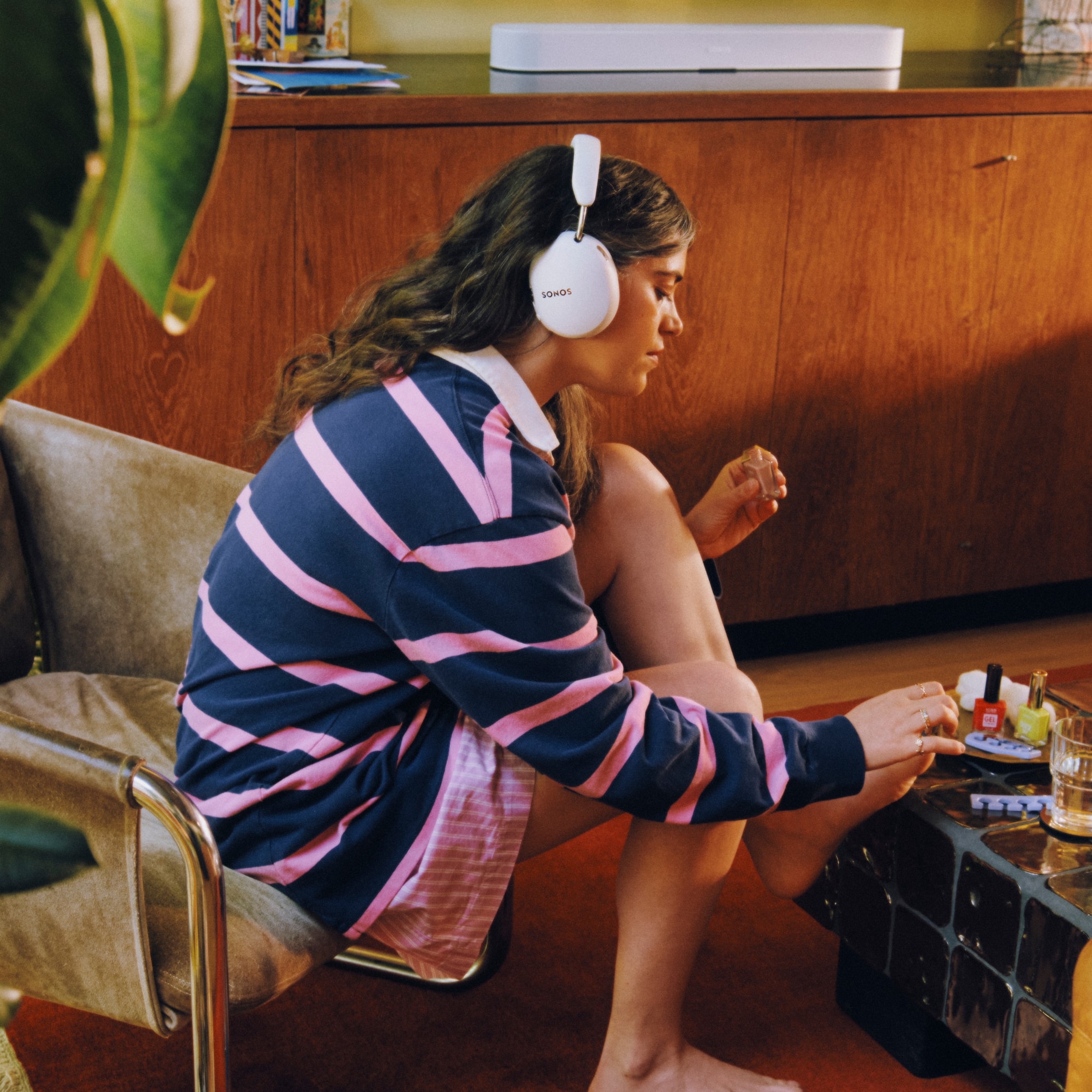 Person wearing white Sonos Ace Ultra headphones sitting in a living room with plants and wooden furniture while painting their nails