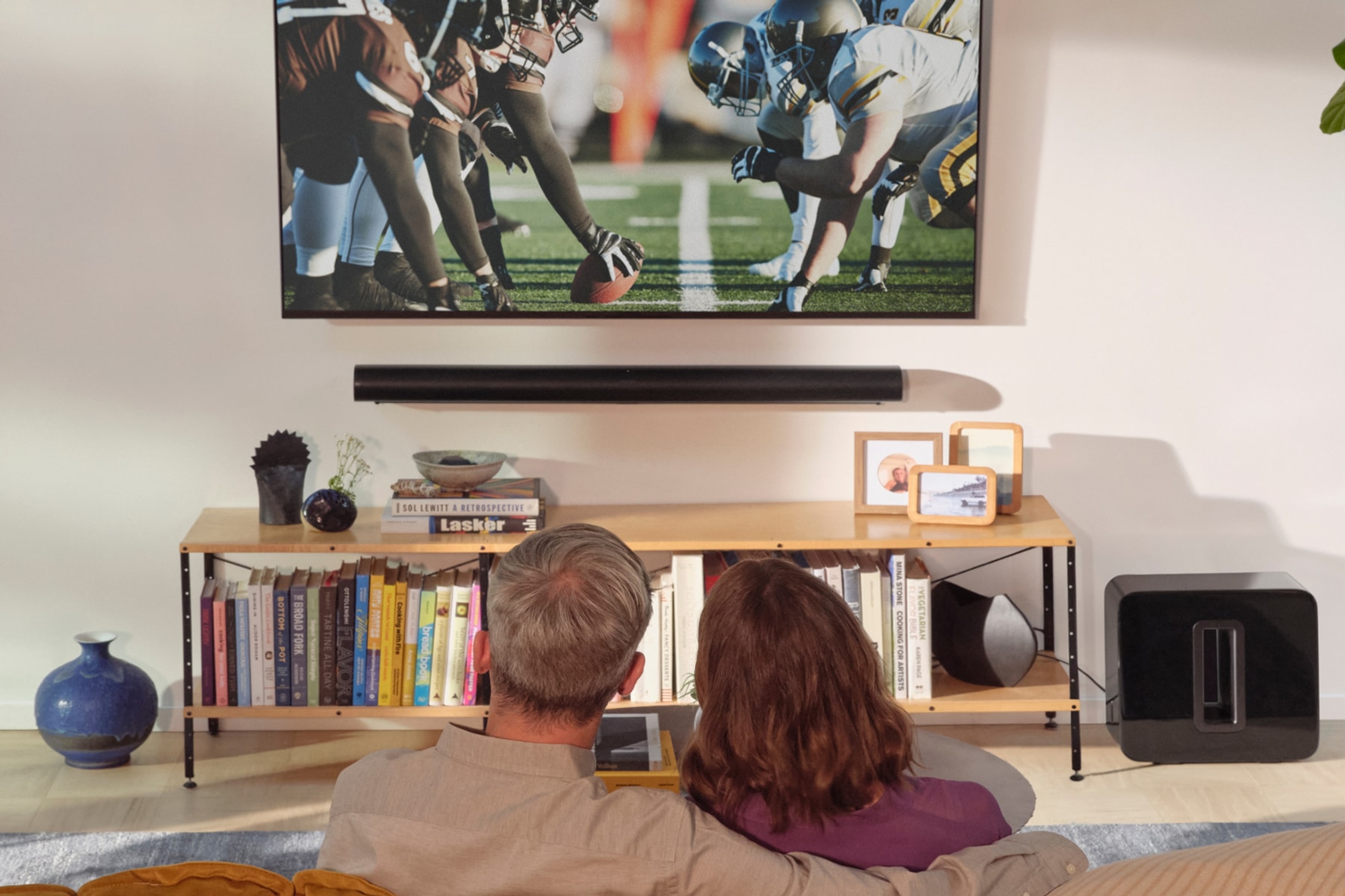 A couple on the couch watching a football game with a black Sonos Arc Ultra and Sub 4