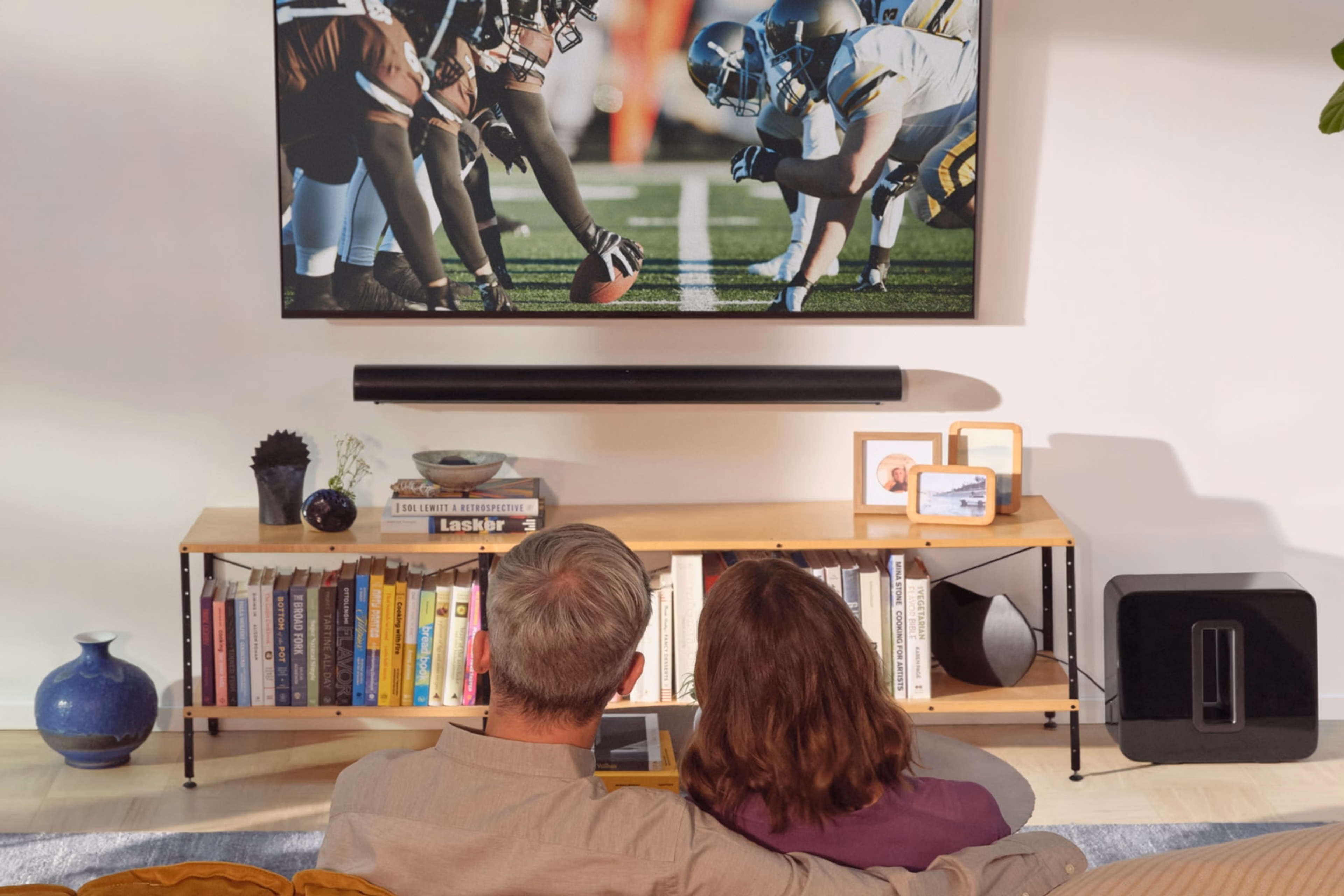 A couple on the couch watching a football game with a black Sonos Arc Ultra and Sub 4