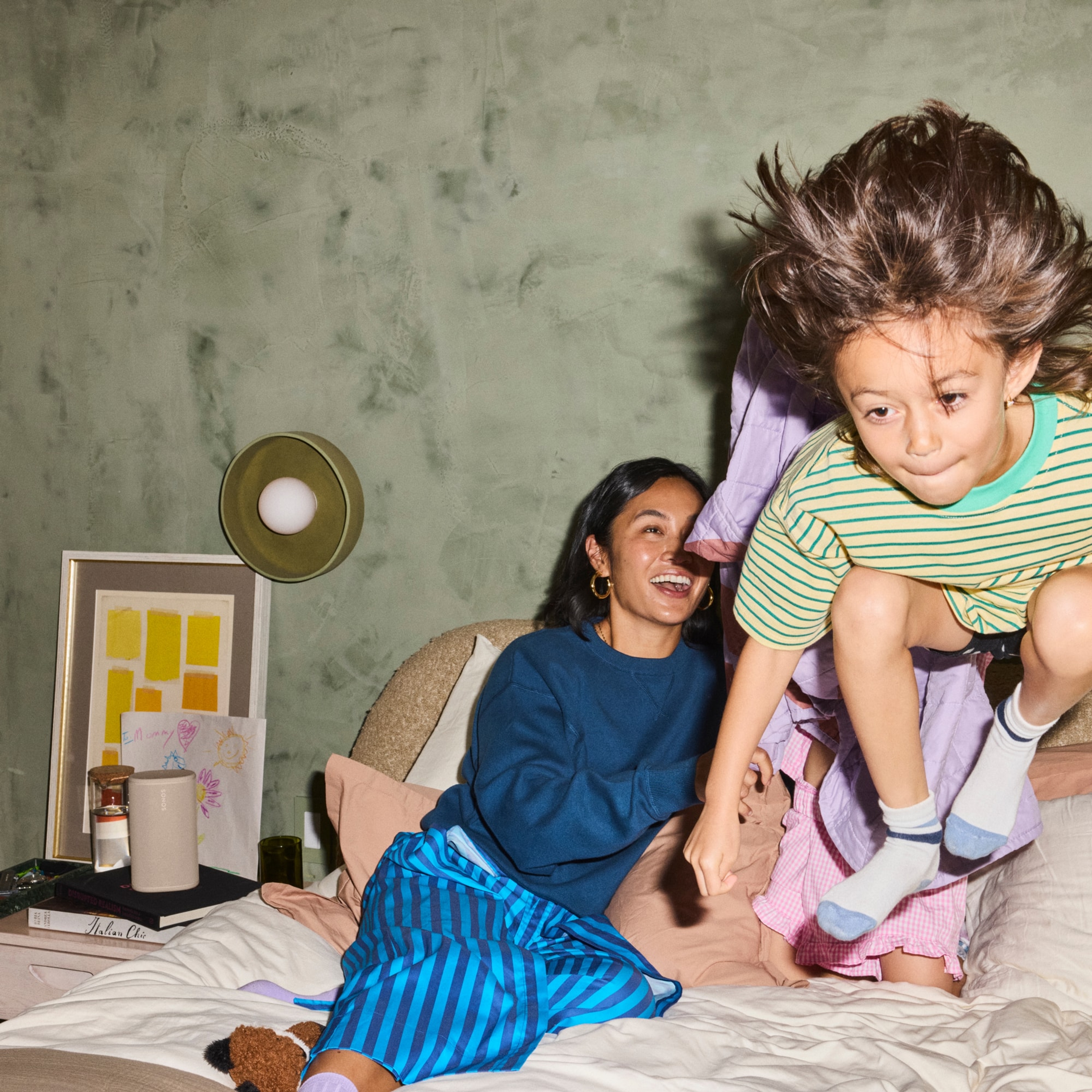 Sonos Play speaker on a night stand with one person sitting and a kid jumping on the bed