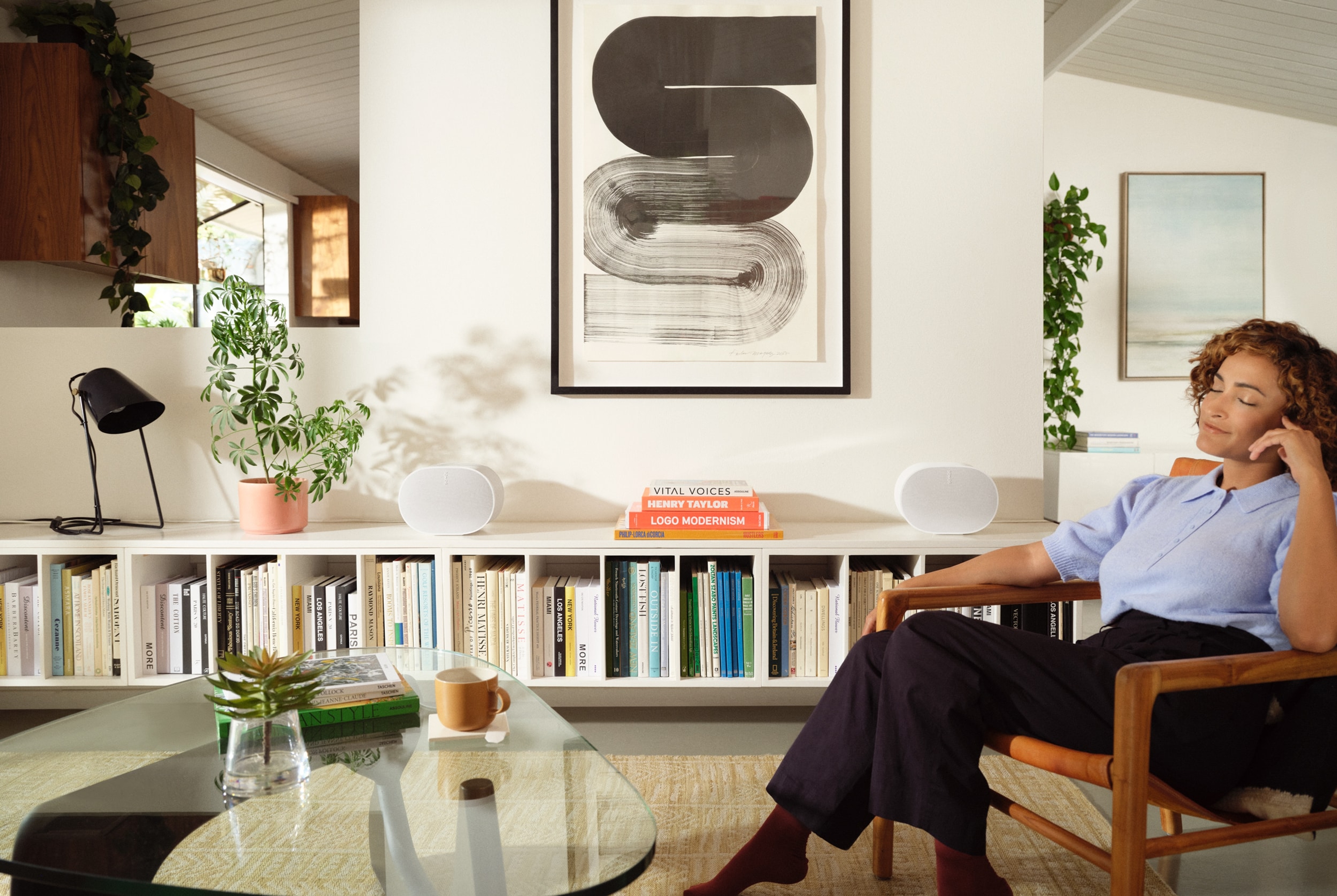 A woman sitting on a wooden chair listening to two white Sonos Era 300 speakers kept on the bookshelf