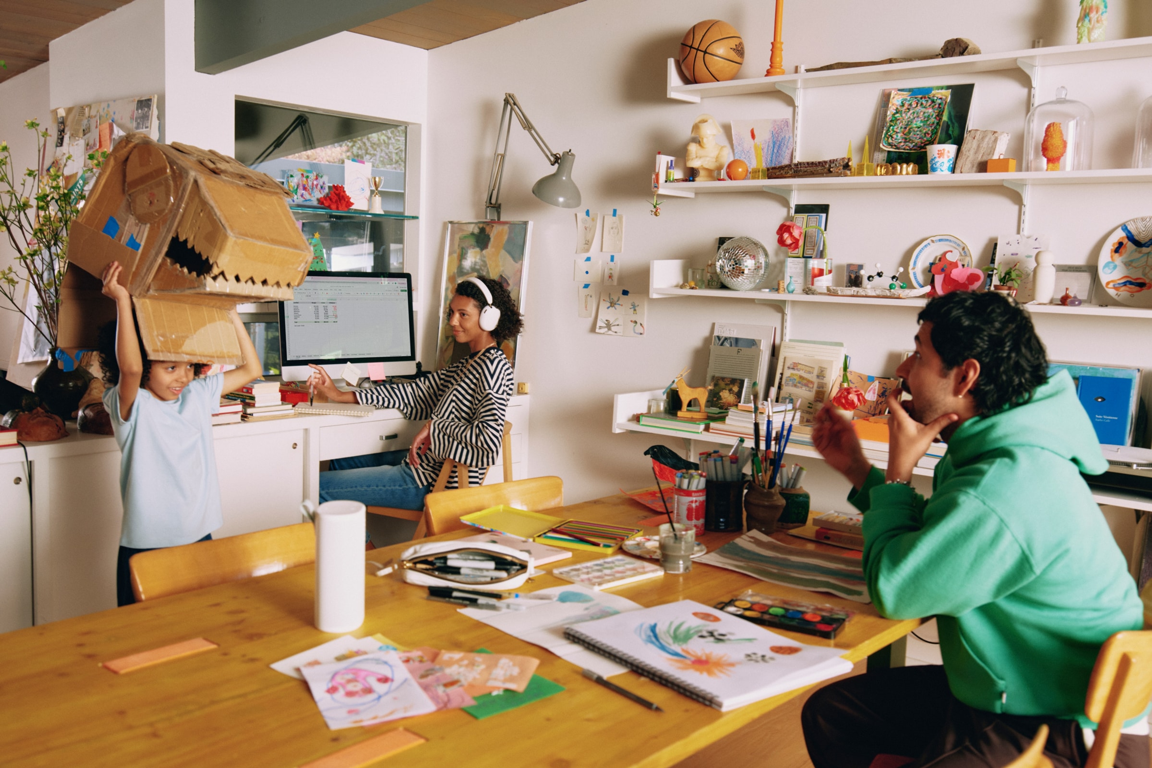 Family at the table making arts and crafts together listening to a white Sonos Play, while mom works at the computer wearing white Sonos Ace Ultra headphones