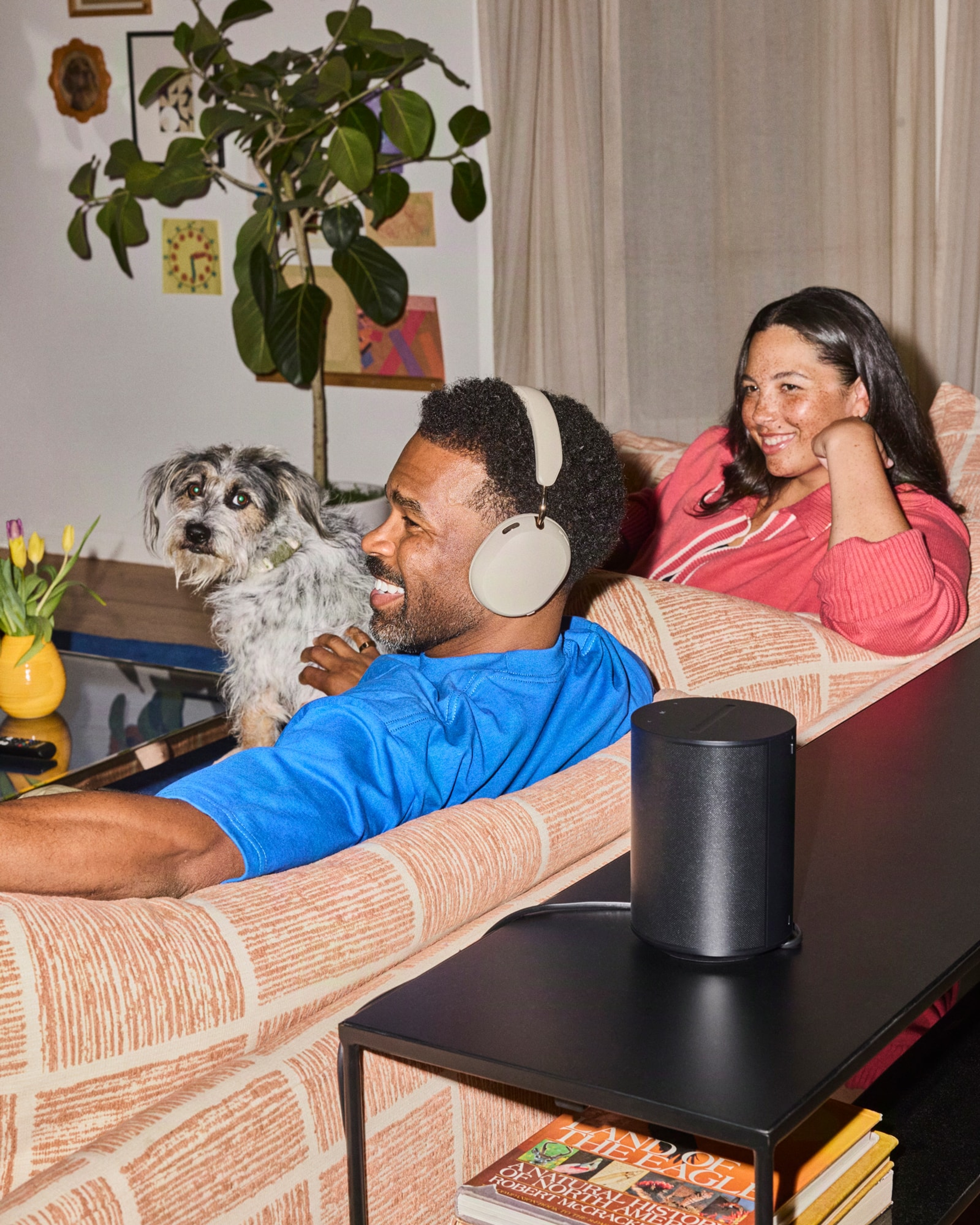 Couple seated on a couch with their dog, one wearing white Sonos Ace Ultra headphones