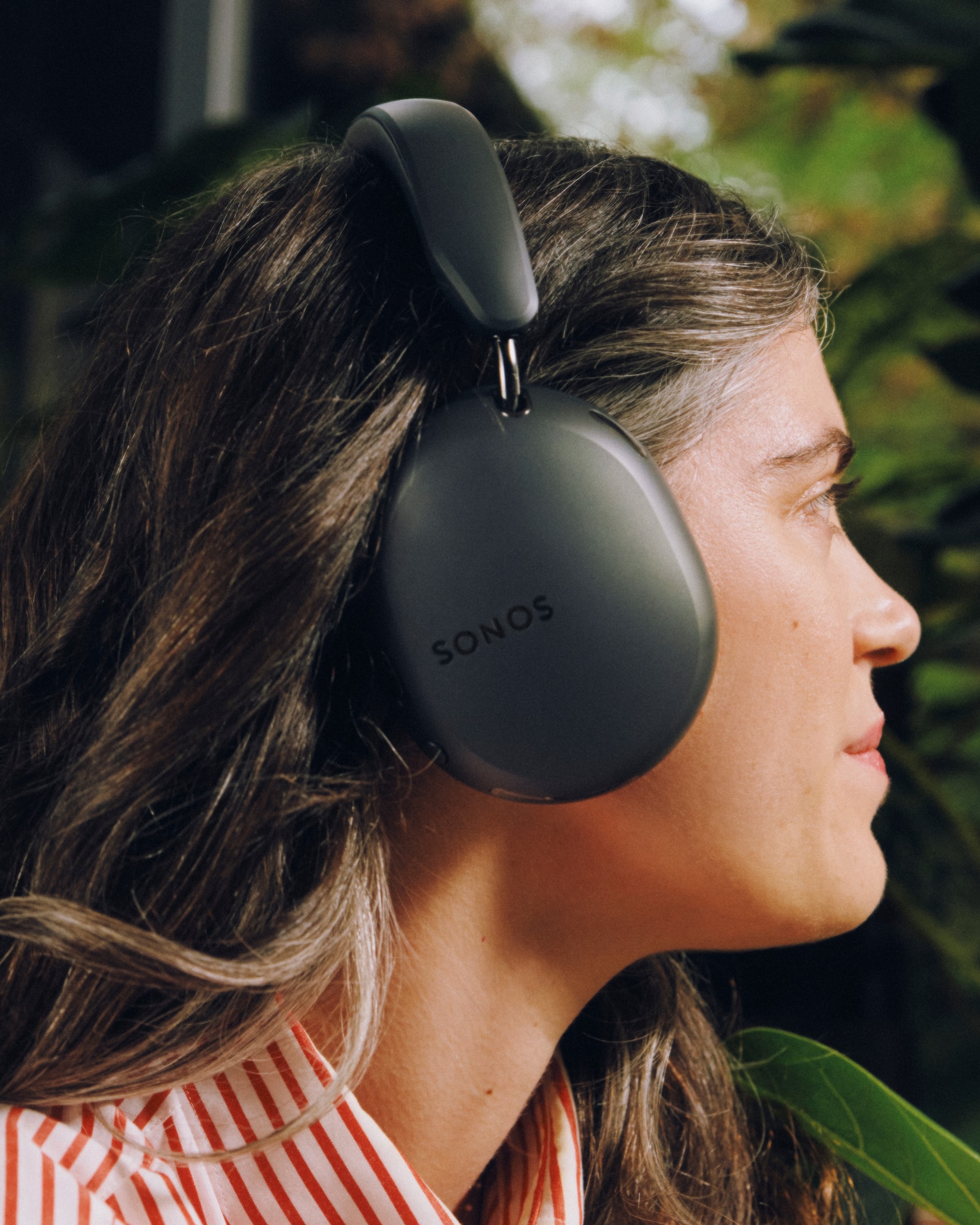 Person in a red striped shirt sitting on a patio among plants and wearing black Sonos Ace Ultra headphones