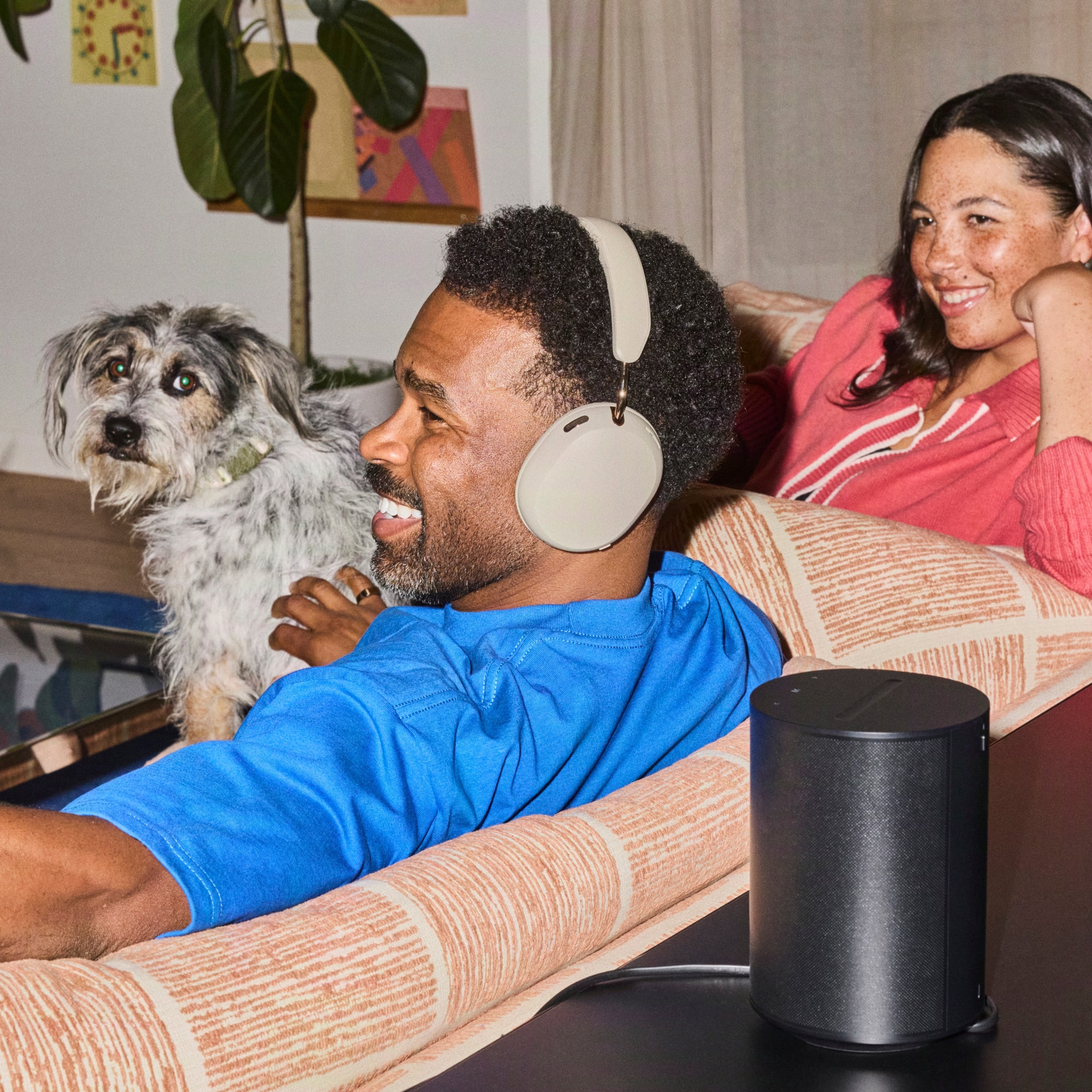 Couple seated on a sofa with their dog, one wearing sand-coloured Sonos Ace Ultra headphones