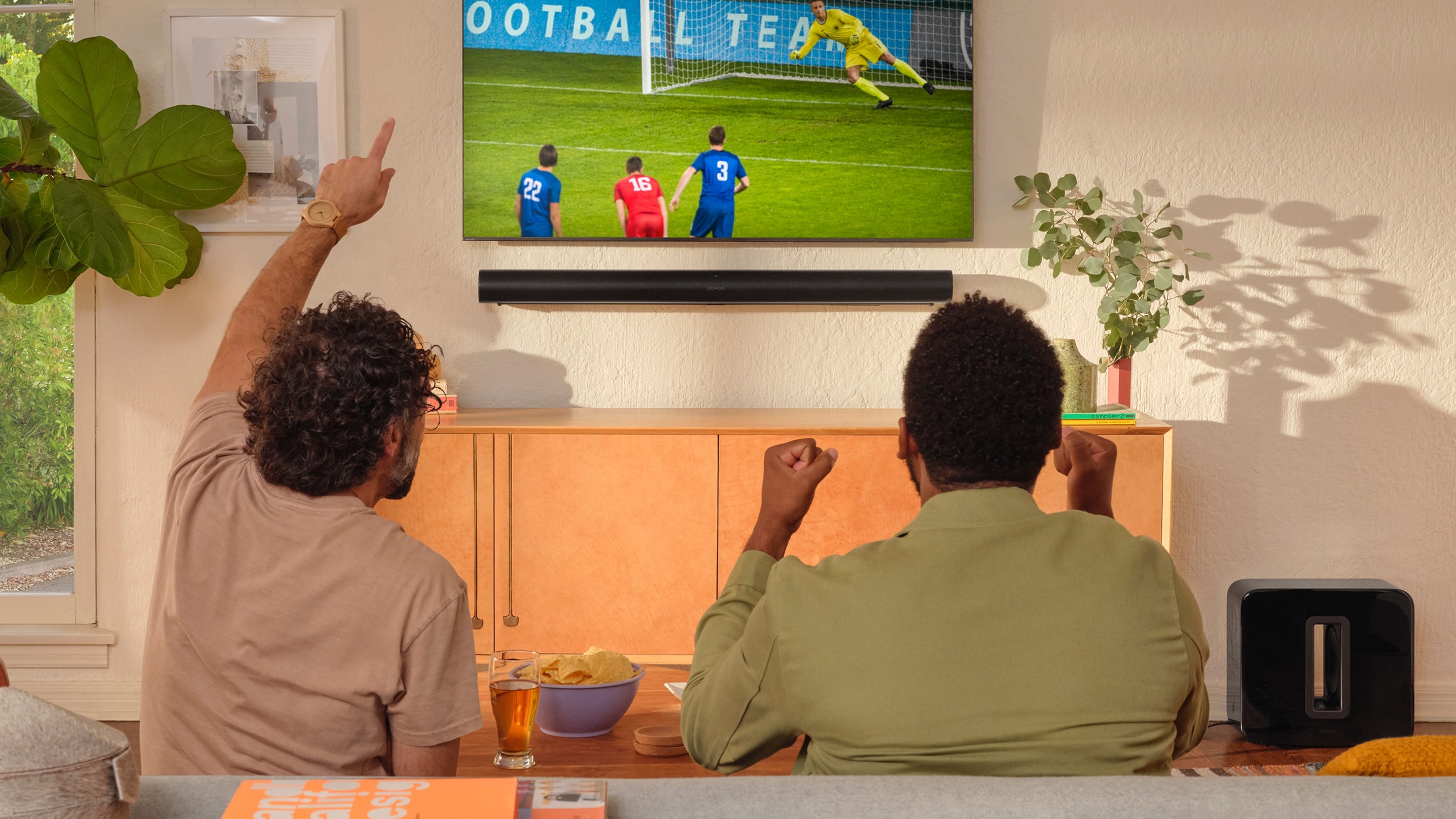 Zwei Personen, die sich auf einem Fernseher mit einer Arc in Schwarz und einem Sub Fußball ansehen