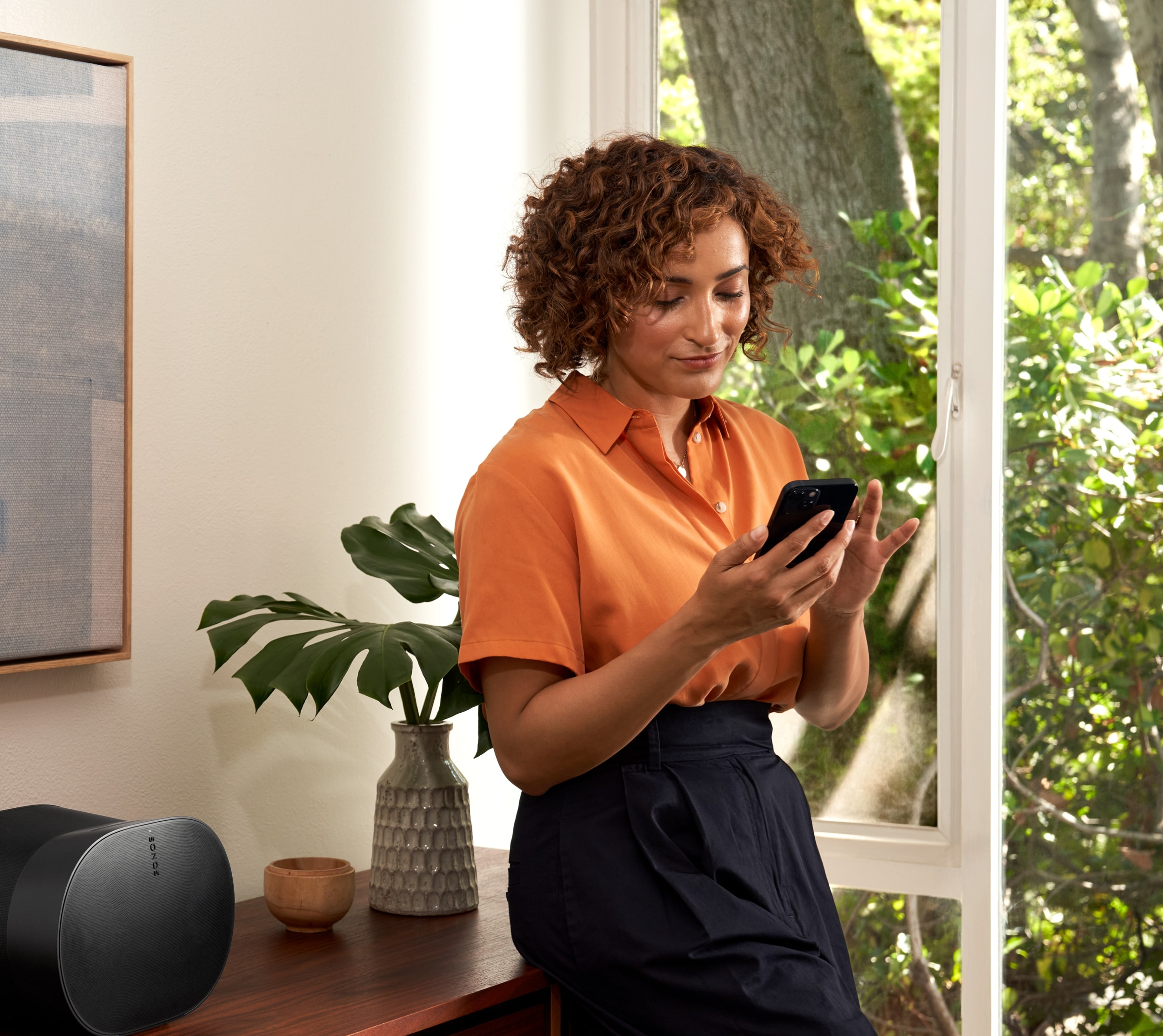 Woman leaning on a credenza using her phone to control a black Sonos Era 300