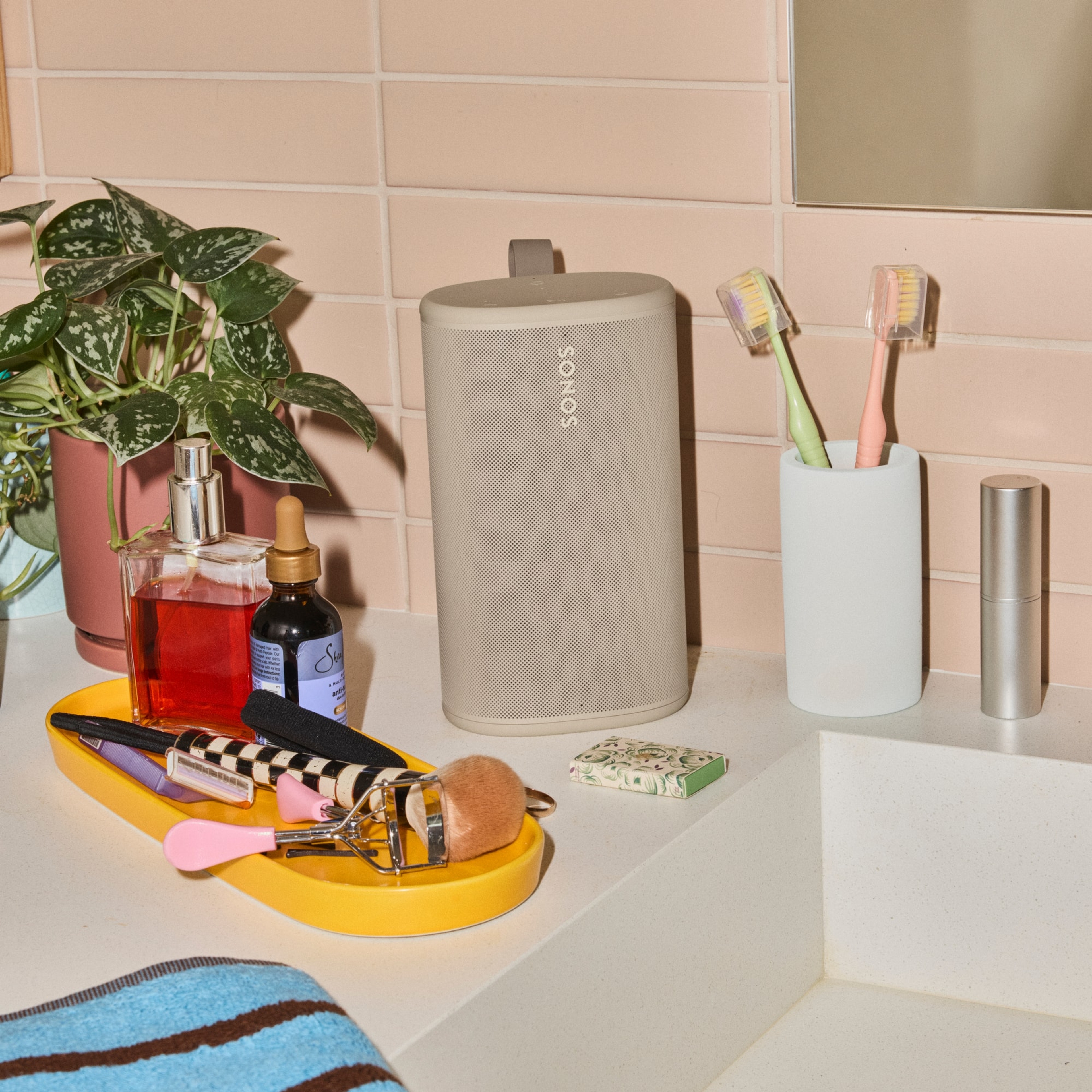 Une enceinte Sonos Play couleur Sable posée sur un comptoir de salle de bain, entourée de produits de beauté et d’articles de toilette