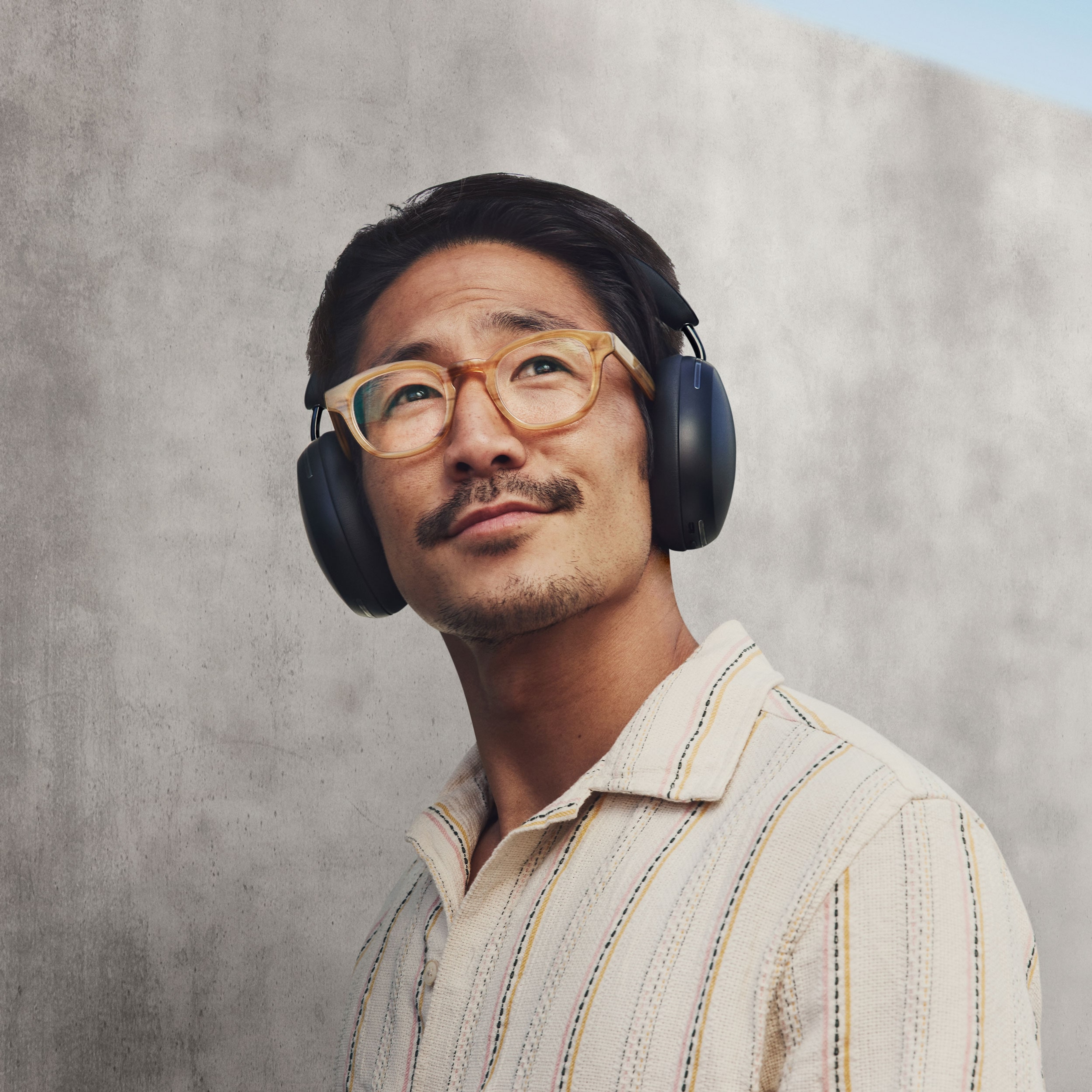 Una persona sonríe y escucha música en sus audífonos Sonos Ace en negro frente a una pared de concreto