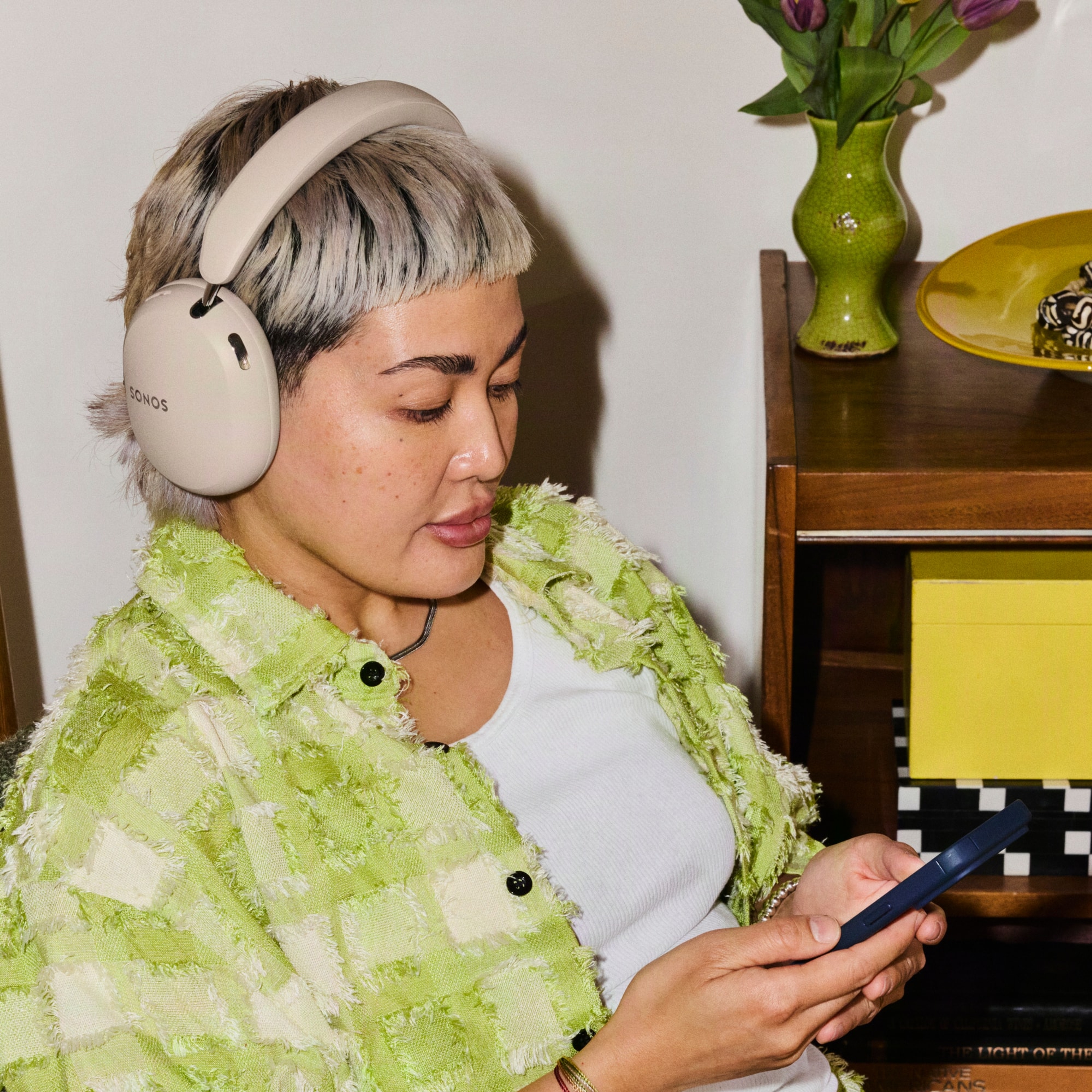 Person wearing sand colored Sonos Ace Ultra headphones and a lime green shirt, holding a phone in a home setting