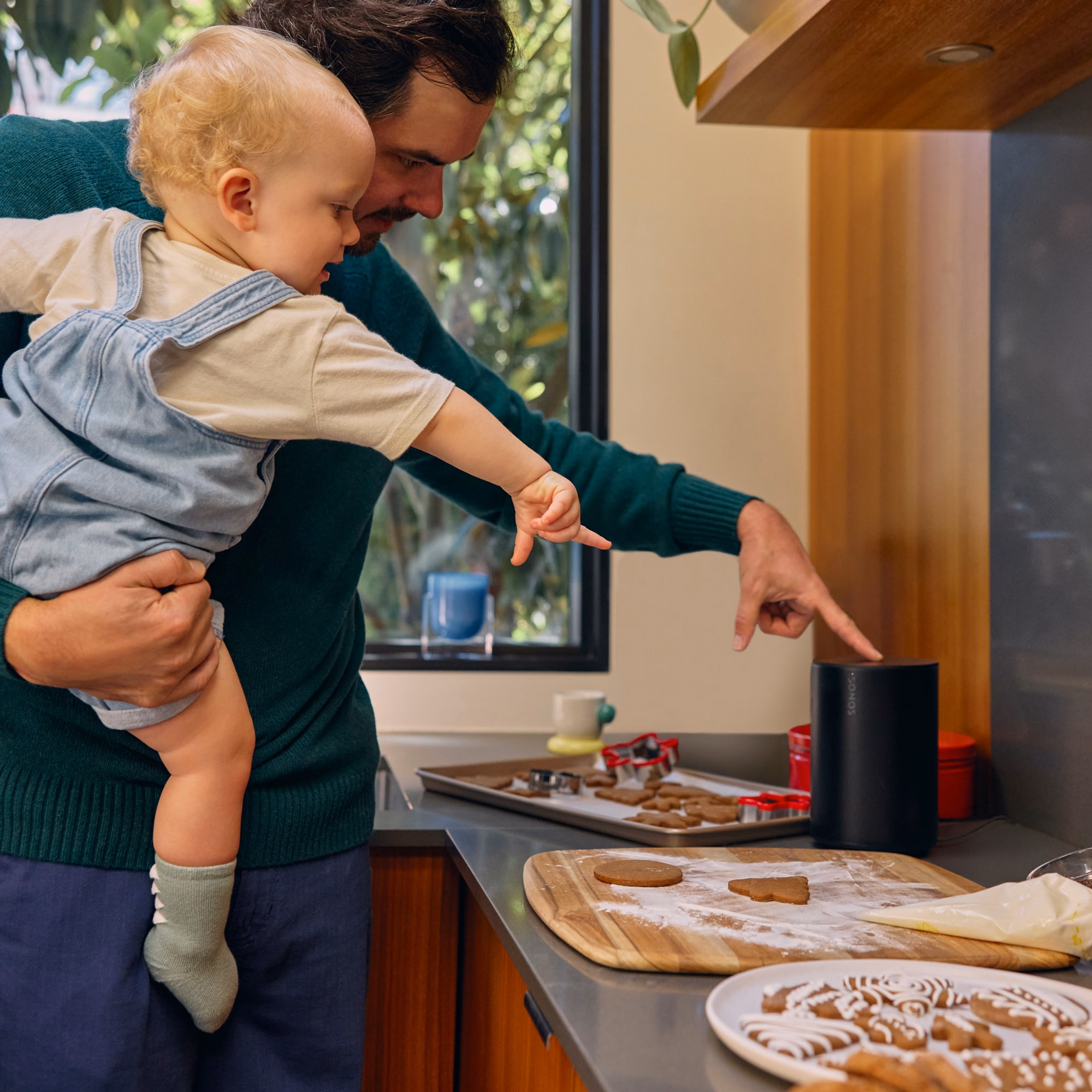 A dad holding his infant while making holiday cookies listening to a black Sonos Era 100 speaker