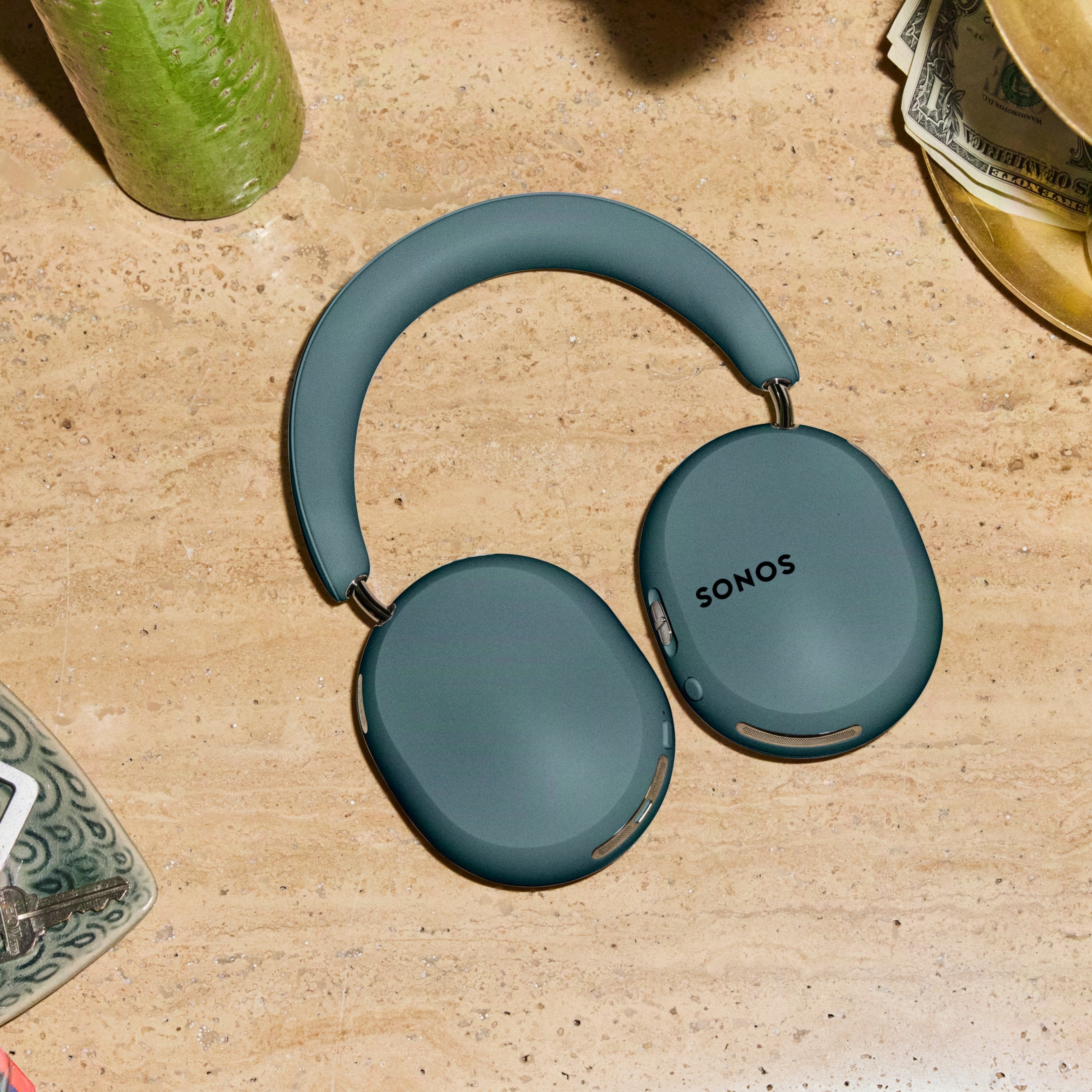 Sonos Ace Ultra headphones in Agave on a wooden tabletop surrounded by plants, sunglasses, and decorative objects