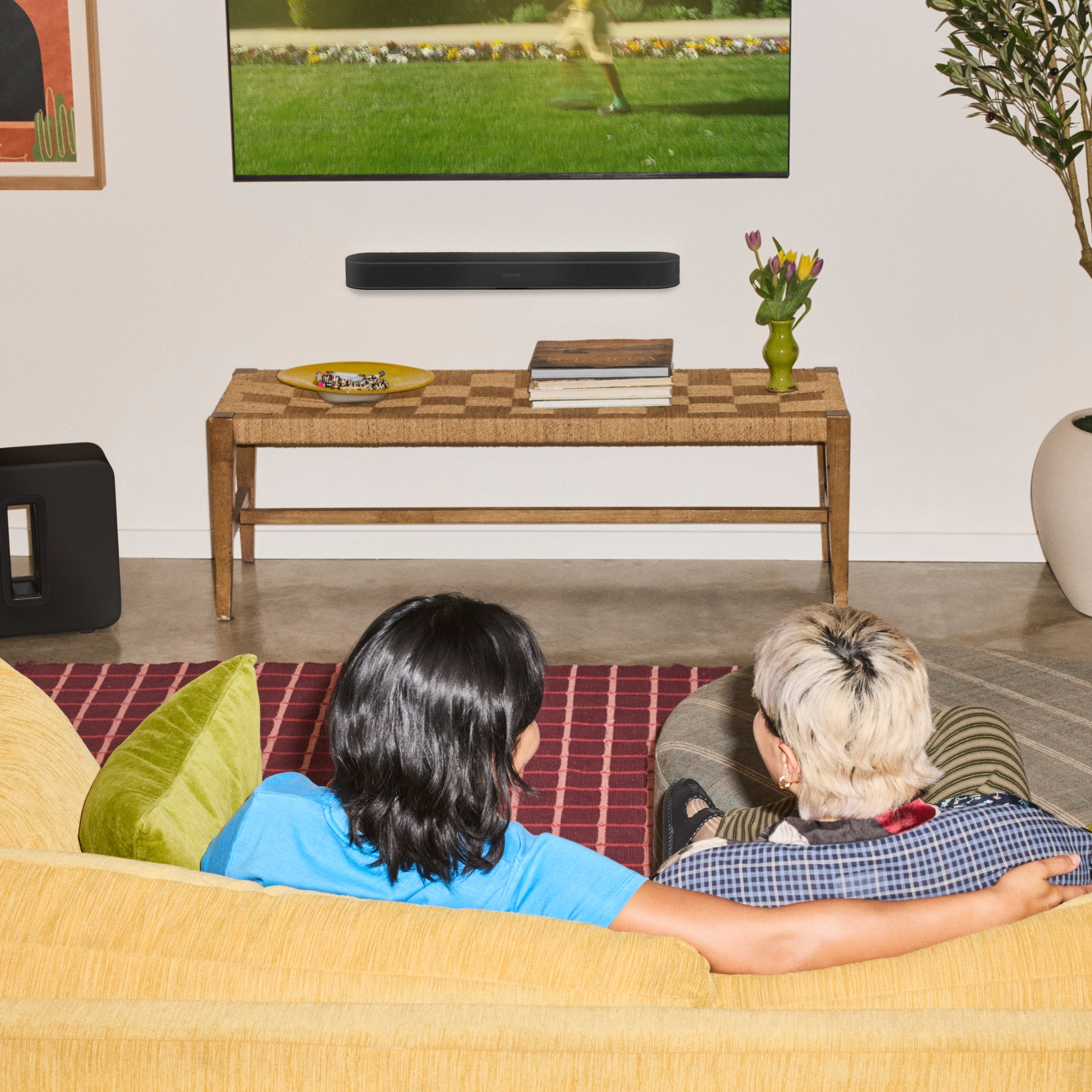 Two people sitting on a yellow couch watching a TV with a black Sonos Beam Ultra soundbar mounted on the wall