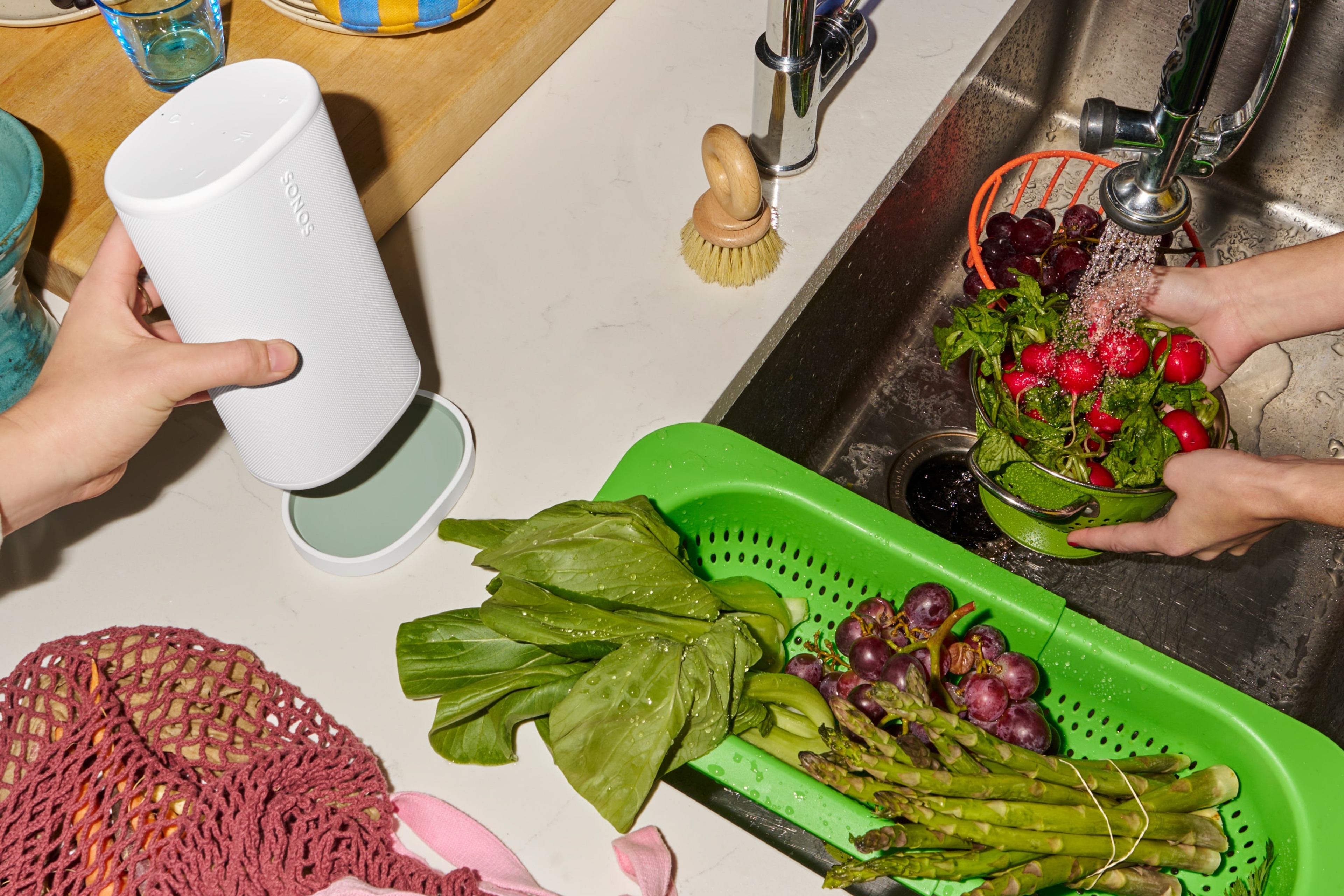 Sonos Playスピーカー（ホワイト）をキッチンシンクの横にある同色の充電ベースに置いている人と、野菜や果物を洗っている人