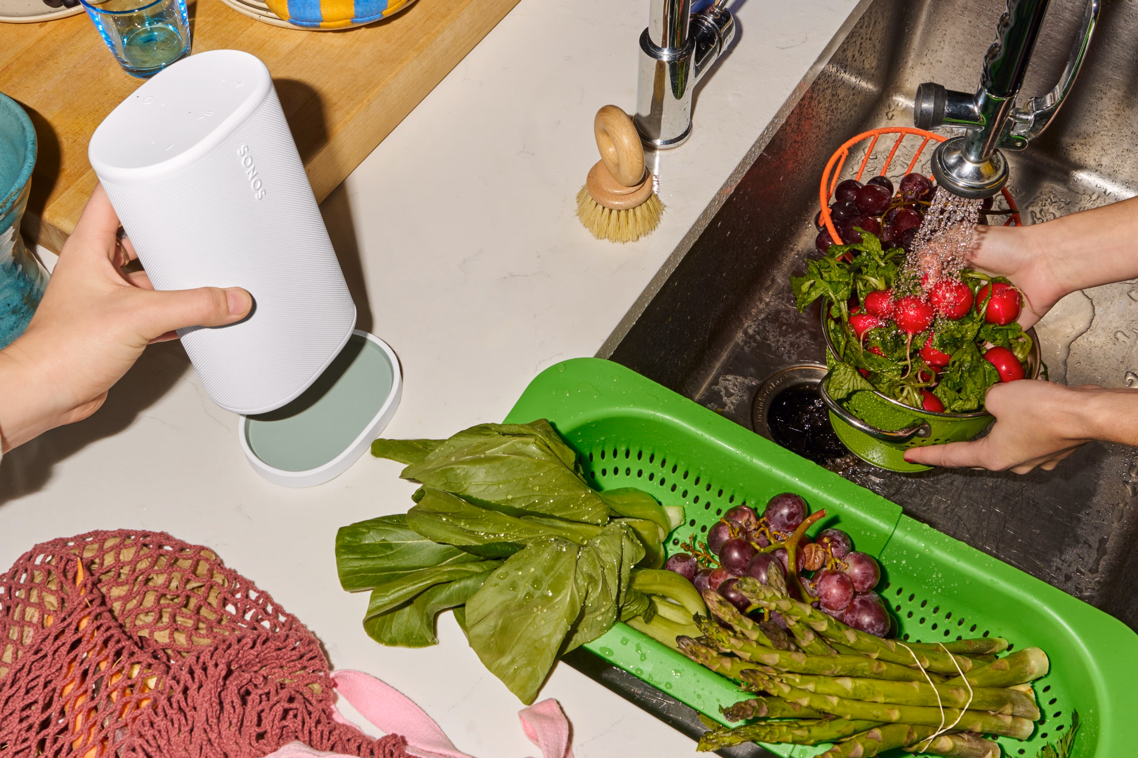 Sonos Playスピーカー(ホワイト)をキッチンシンクの横にある同色の充電ベースに置いている人と、野菜や果物を洗っている人