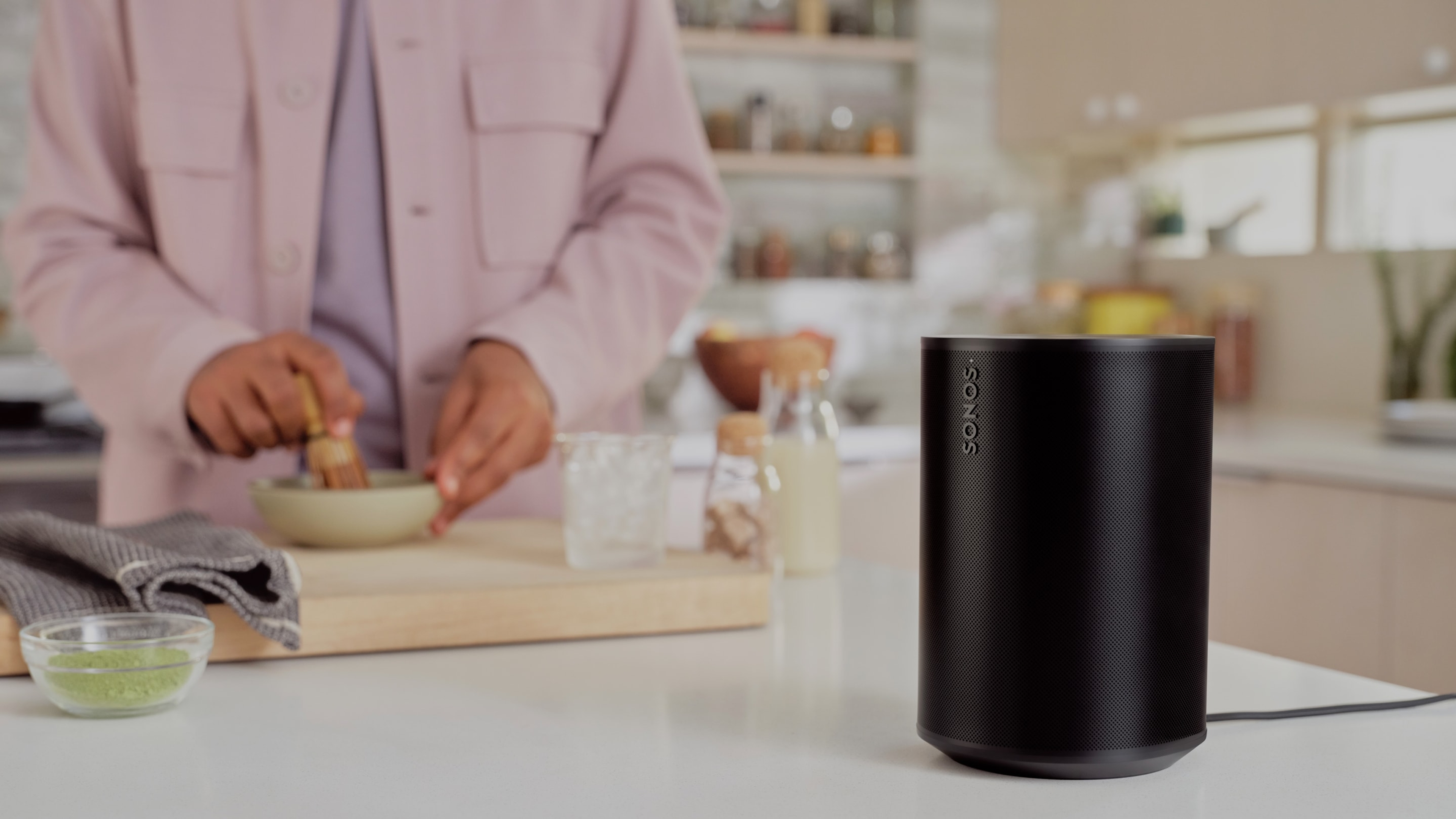 Closeup of a black Era 100 speaker on a kitchen counter while someone makes coffee in the background