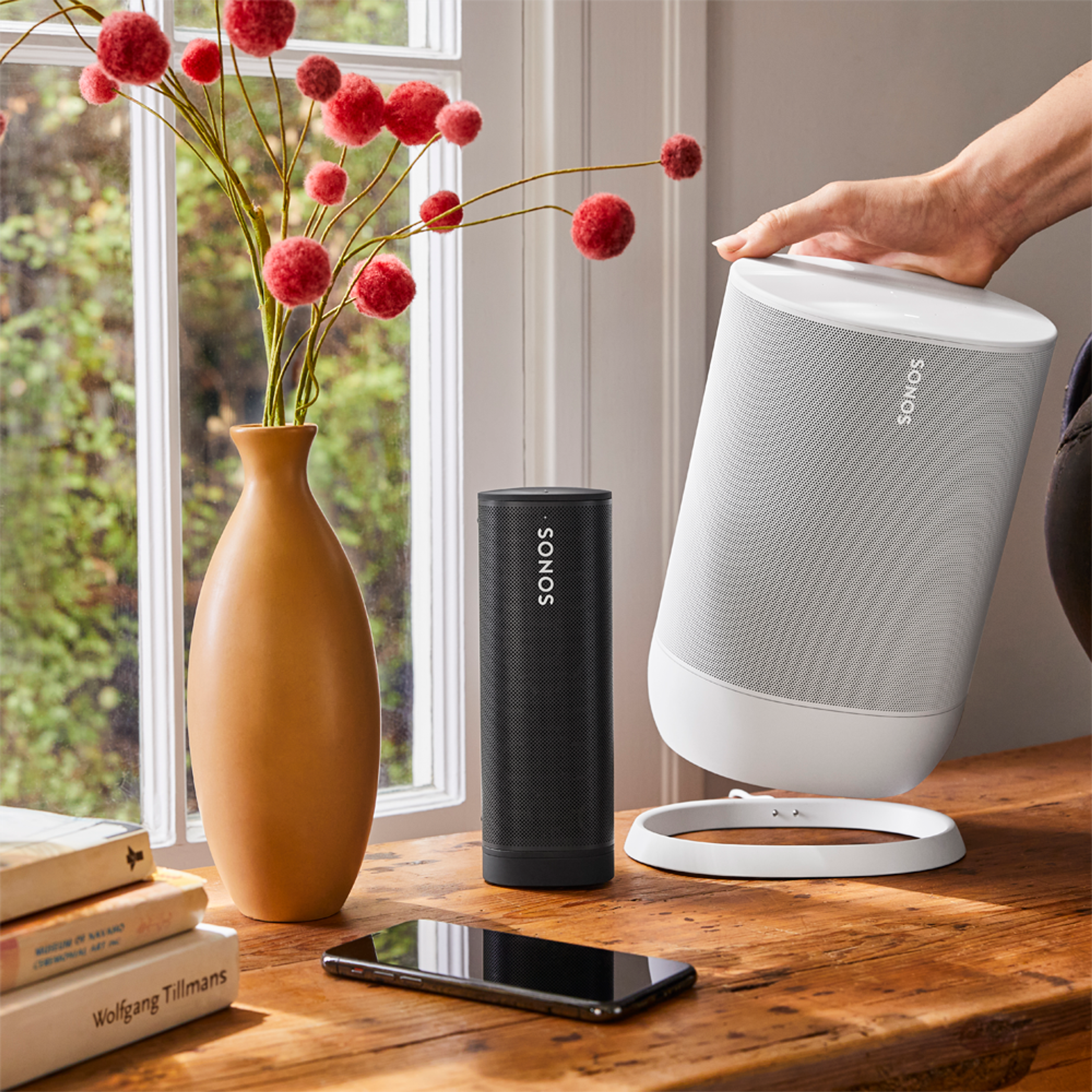 A Sonos Roam resting on a table next to a person's hand picking up a Sonos Move