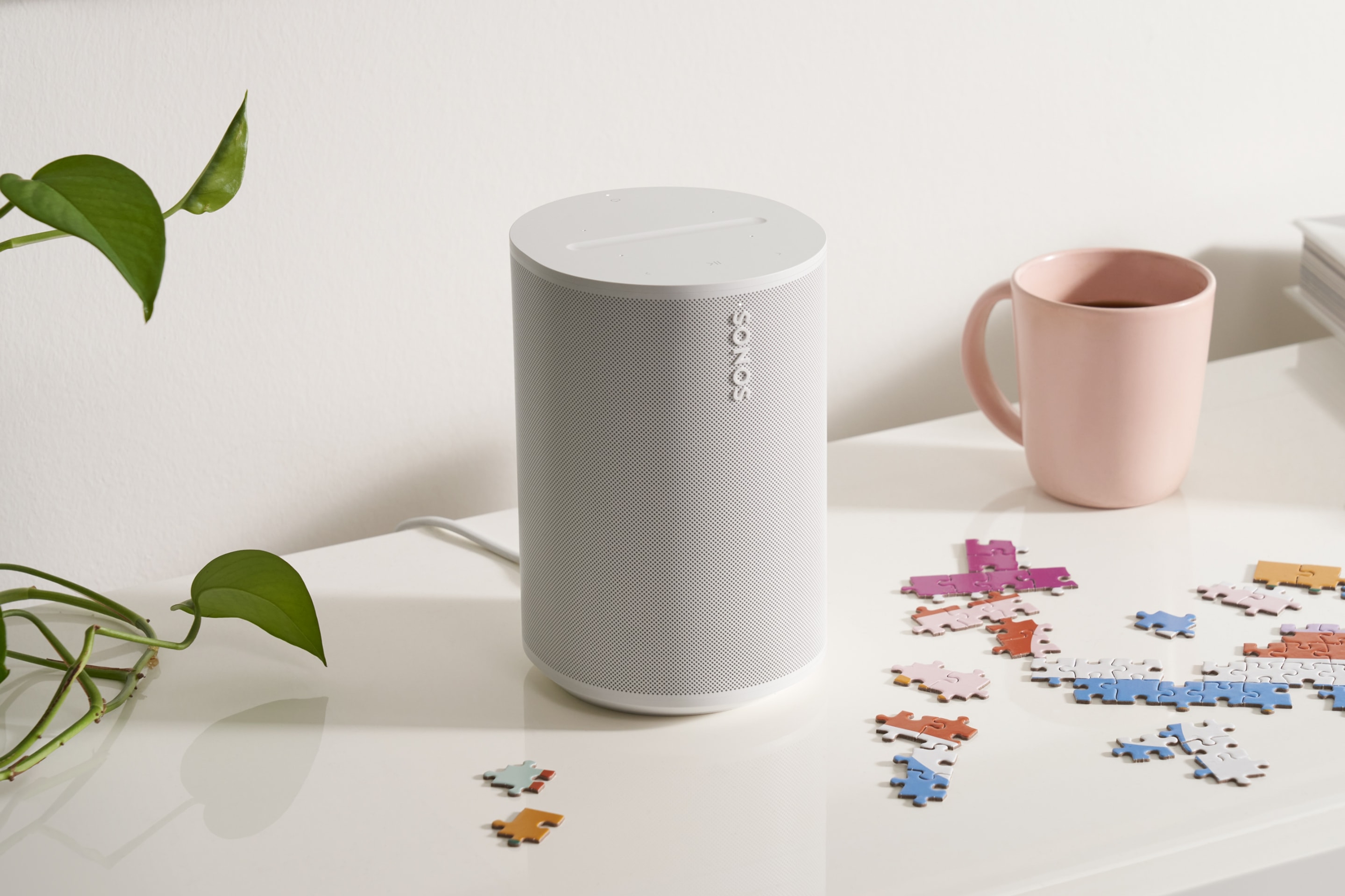 A white Sonos Era 100 on a counter top next to a plant and a coffee mug