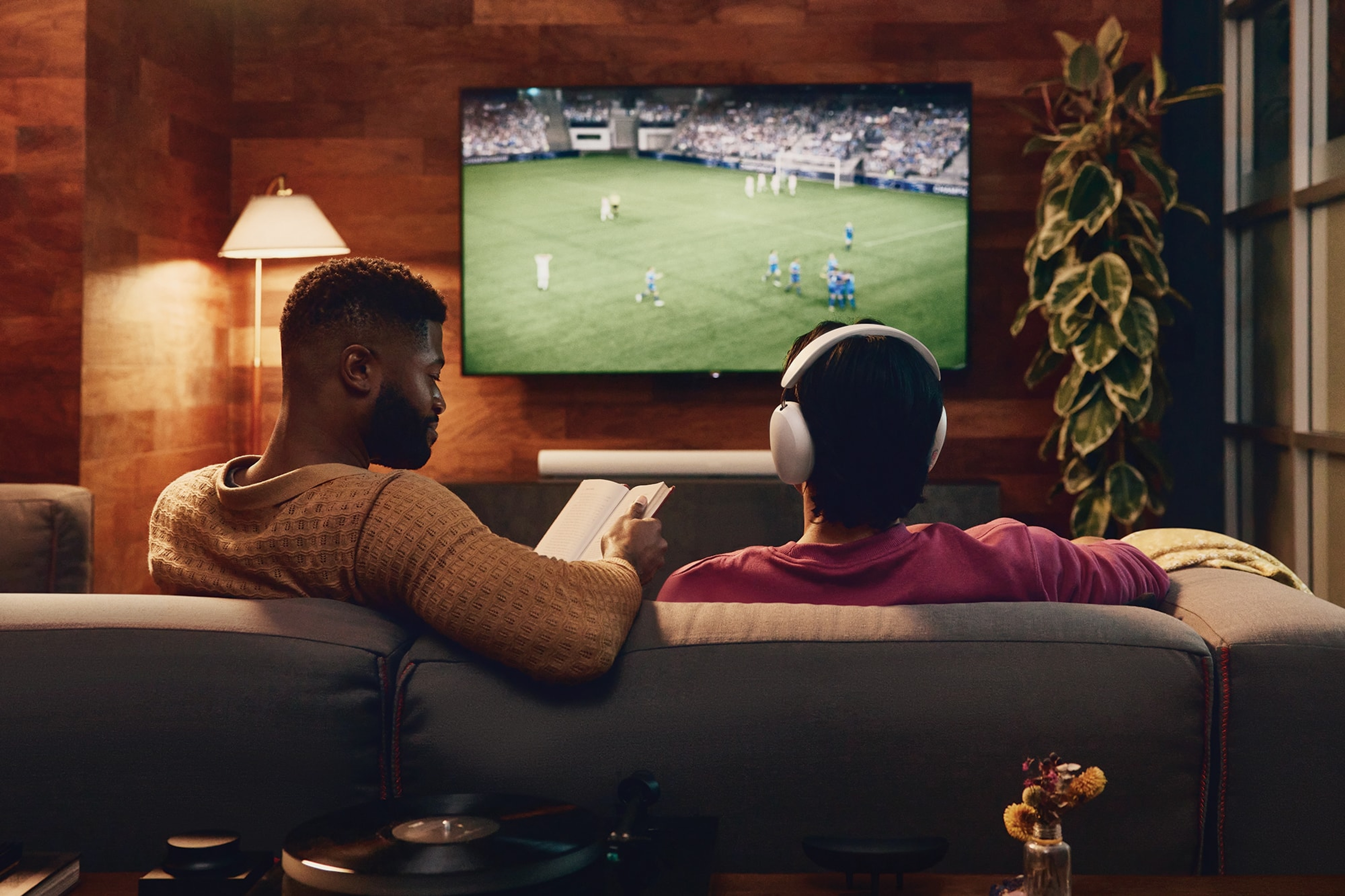 Mensen kijken tv met een witte Arc Ultra; de ene leest, de andere draagt een witte Sonos Ace koptelefoon