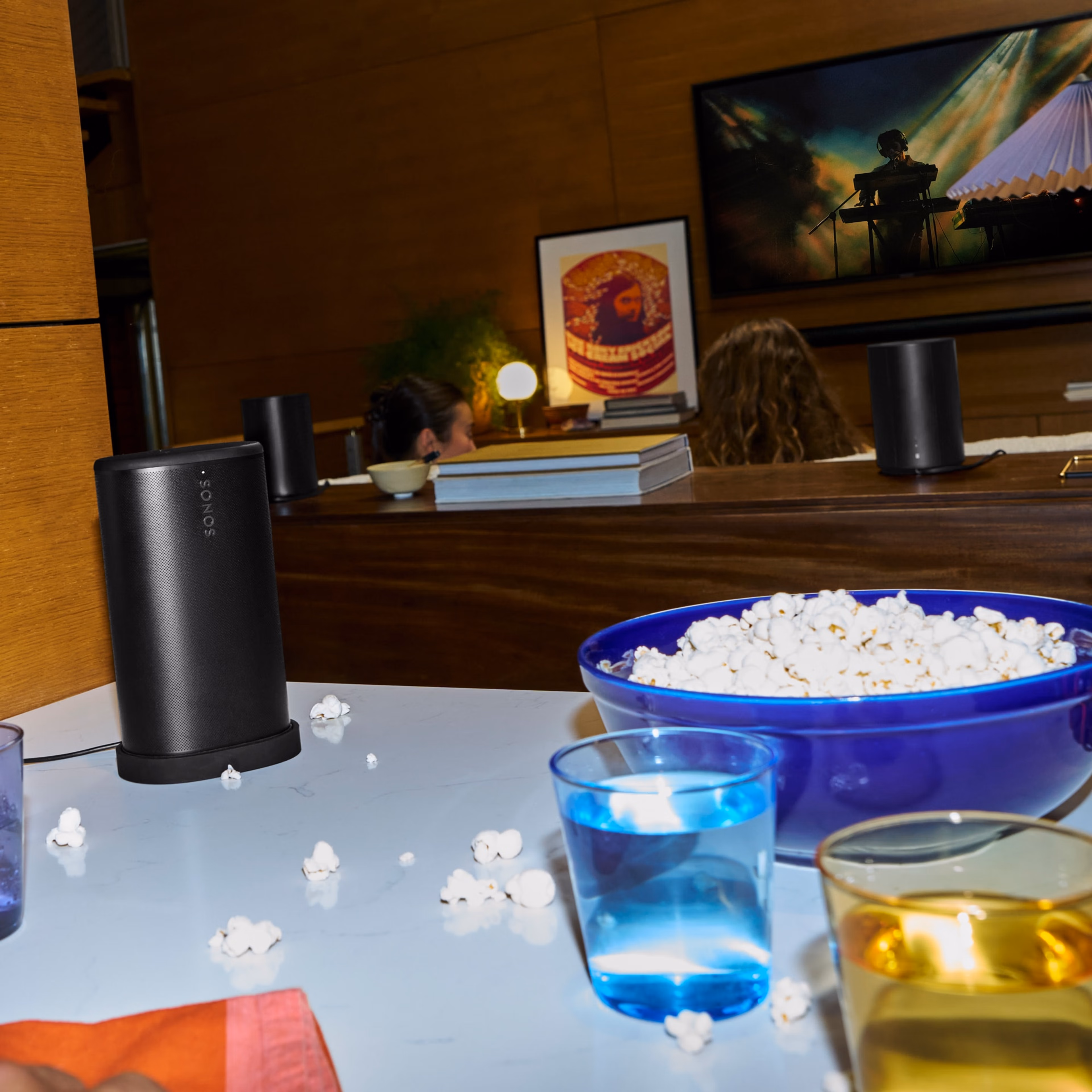 キッチンカウンターにあるポップコーンのボウルの横に置かれたSonos Playスピーカー(ブラック)と、映画を観ている家族
