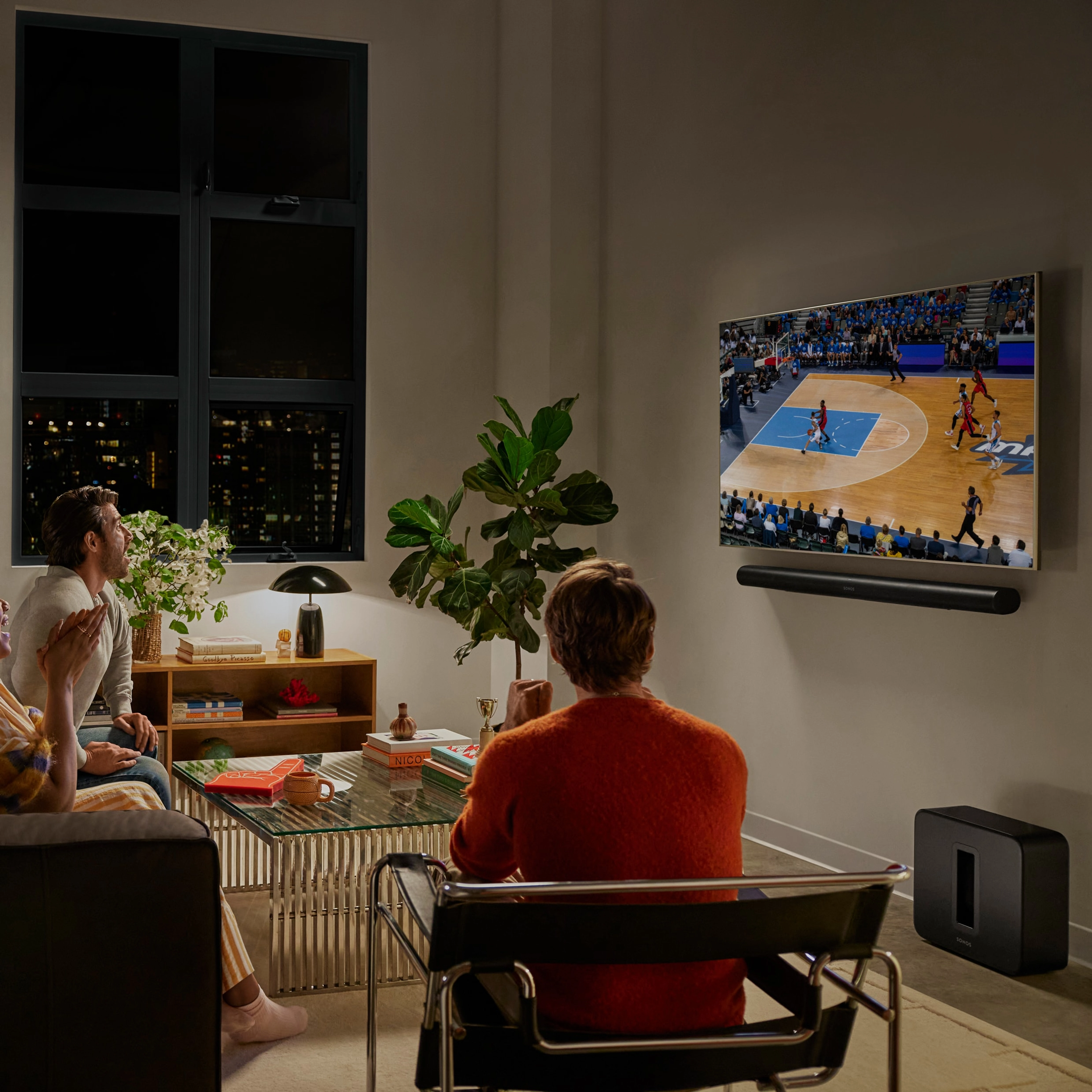 En grupp vänner som tittar på en basketmatch i ett vardagsrum med en svart Arc Ultra och en svart Sub 4