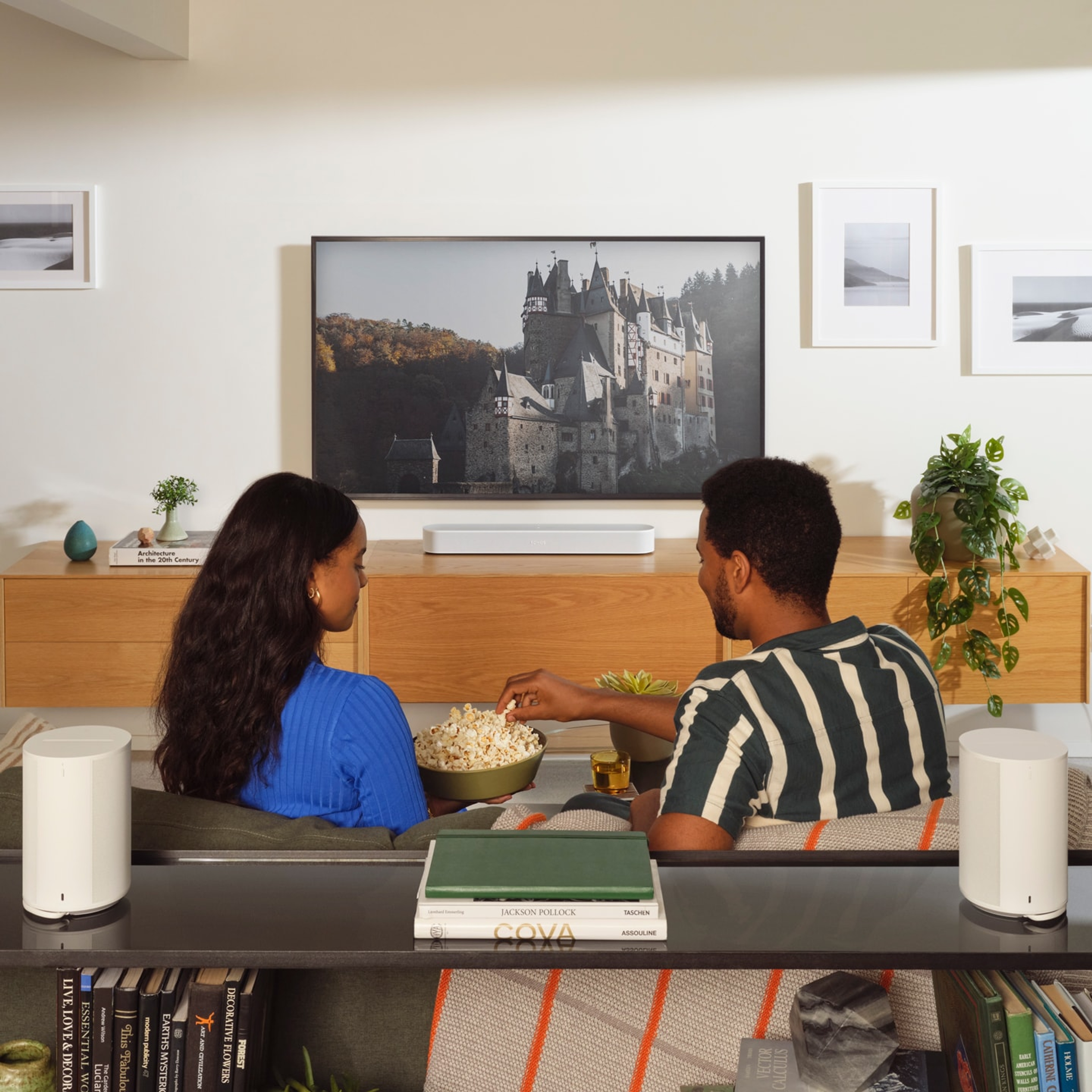 Ein Paar sitzt und isst Popcorn, während es fernschaut, auf einer weißen Sonos Beam Soundbar mit Era 100 Surroundlautsprechern