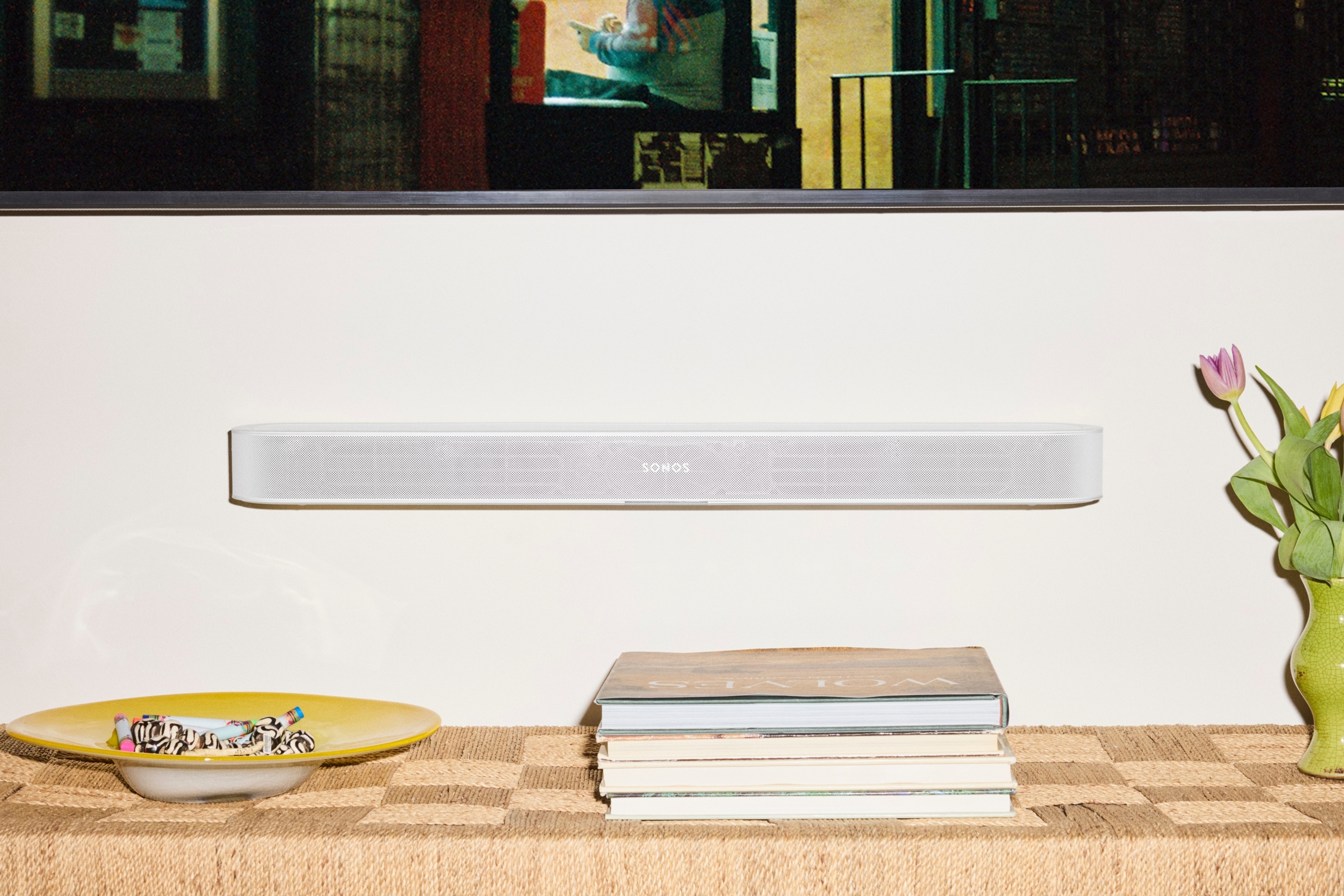 A white Sonos Beam Ultra soundbar mounted on wall above wooden shelf with books and flowers