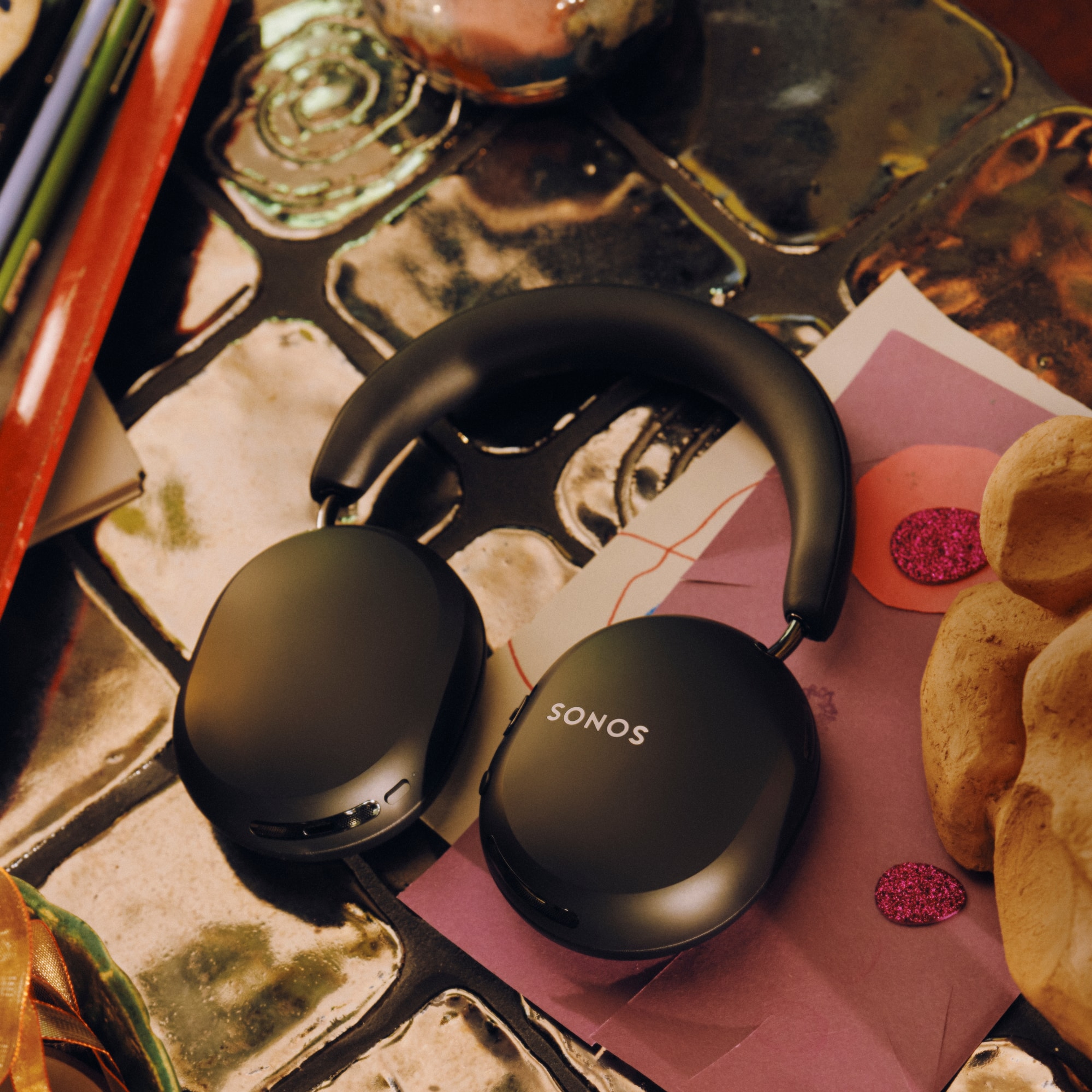 Black Sonos Ace Ultra headphones resting on a pink notebook surrounded by snacks and beverages on a patterned table