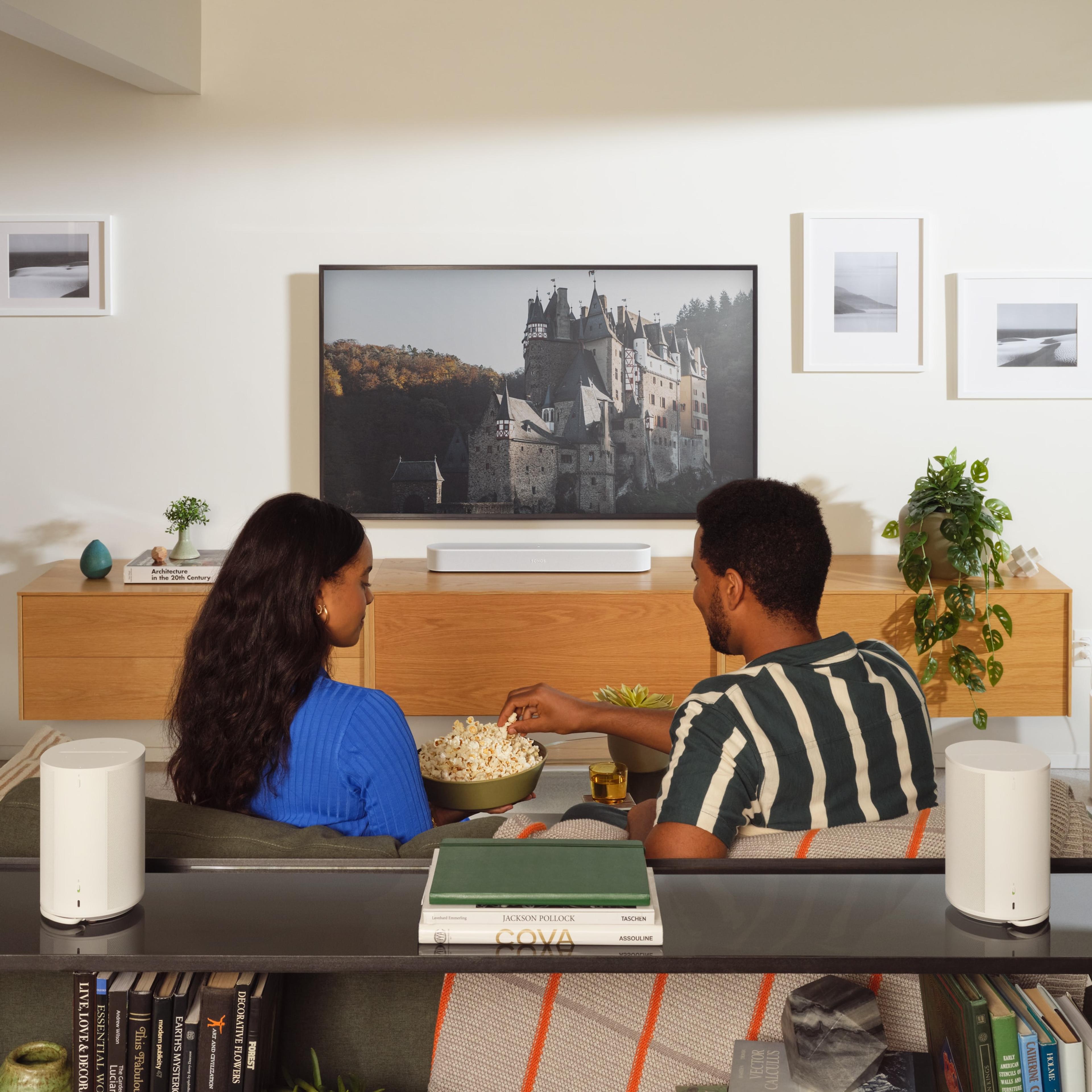 A couple sitting on the couch listening to two white Sonos Era 100 speakers