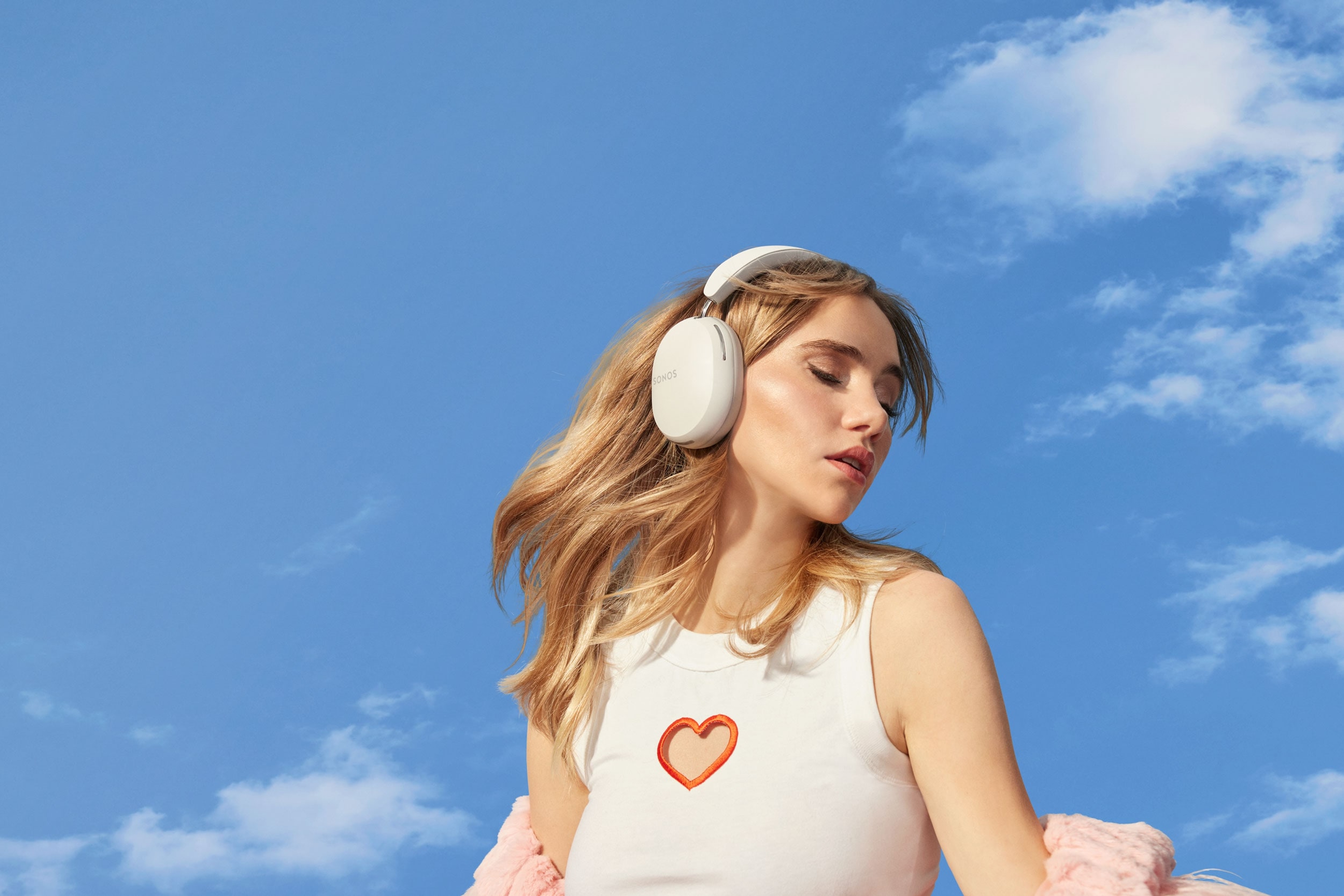 Suki Waterhouse listening to music on soft white Sonos Ace with a blue sky background