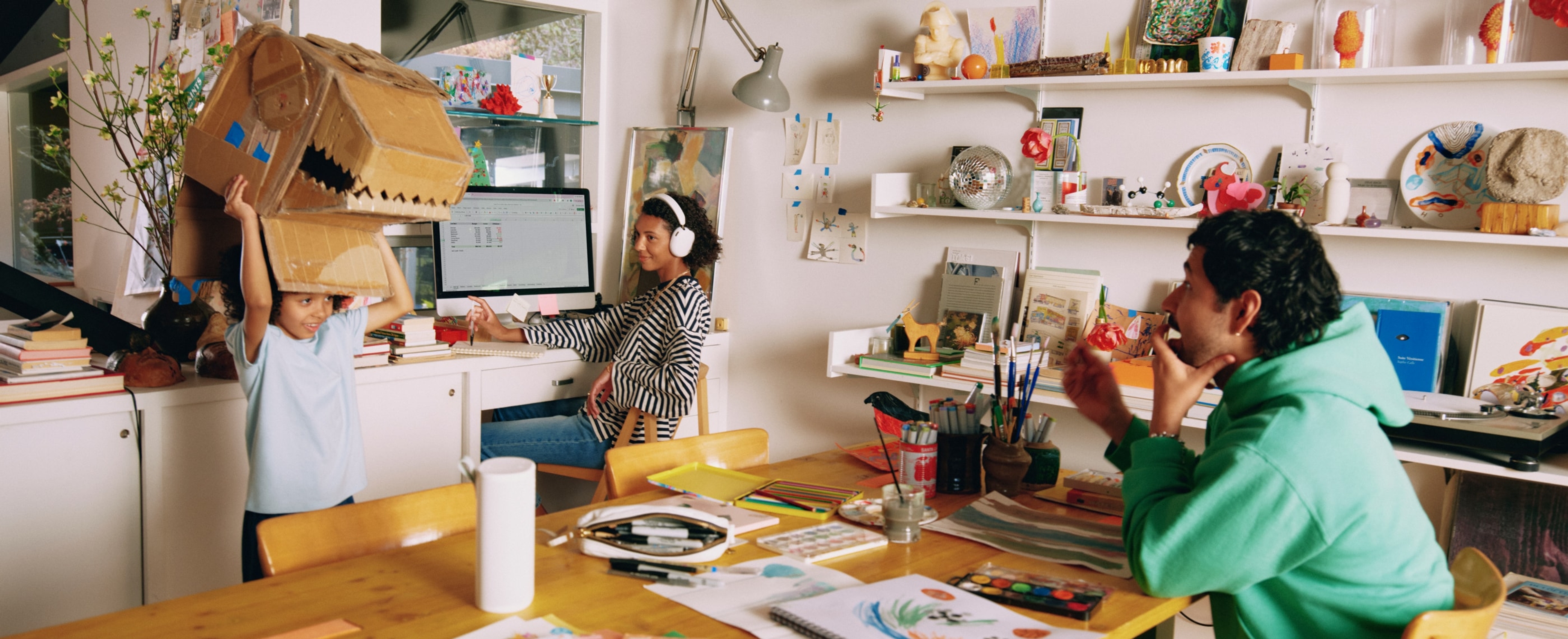 Family at the table making arts and crafts together listening to a white Sonos Play, while mom works at the computer wearing white Sonos Ace Ultra headphones