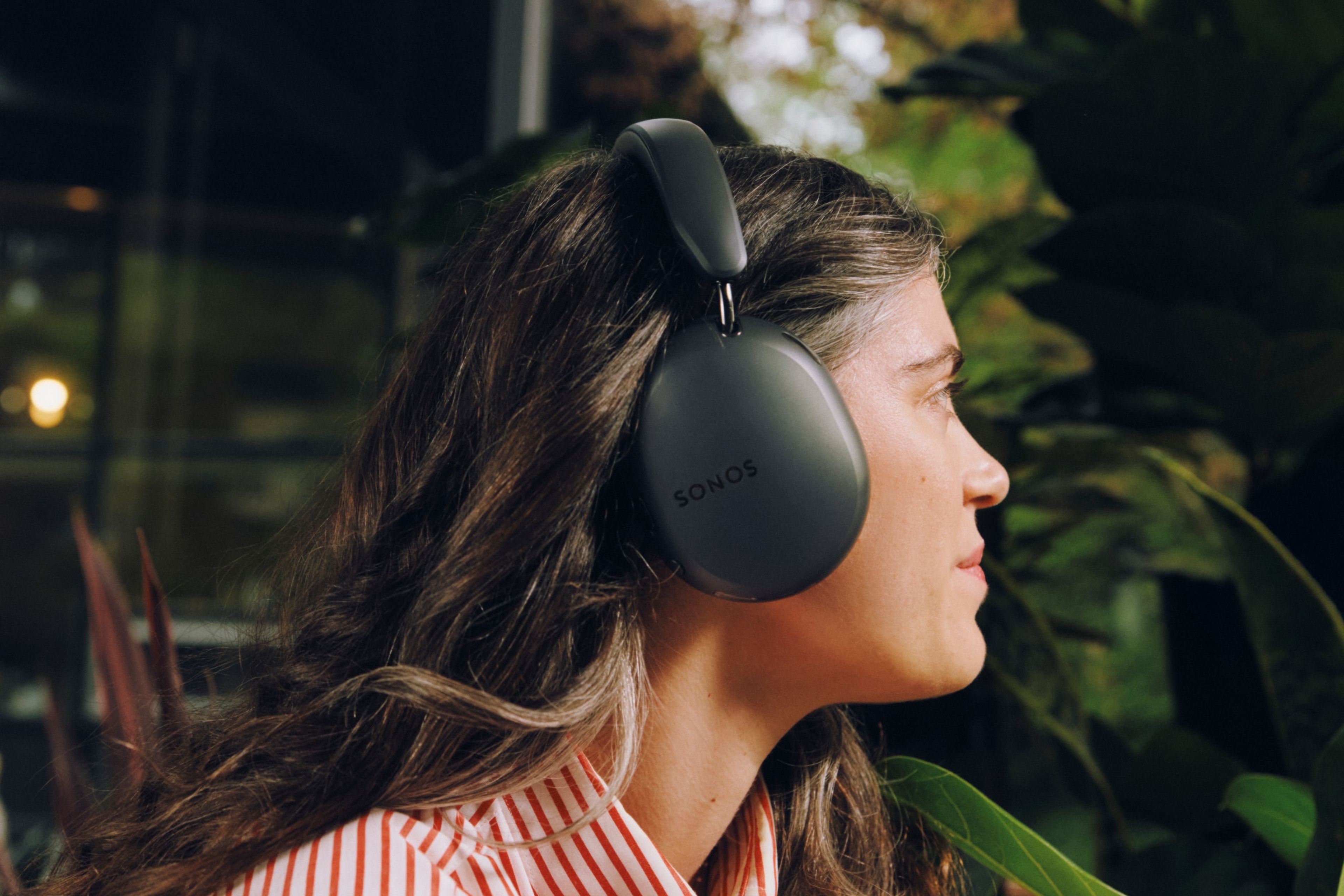 Person wearing black Sonos Ace Ultra headphones in a red striped shirt on a patio among plants