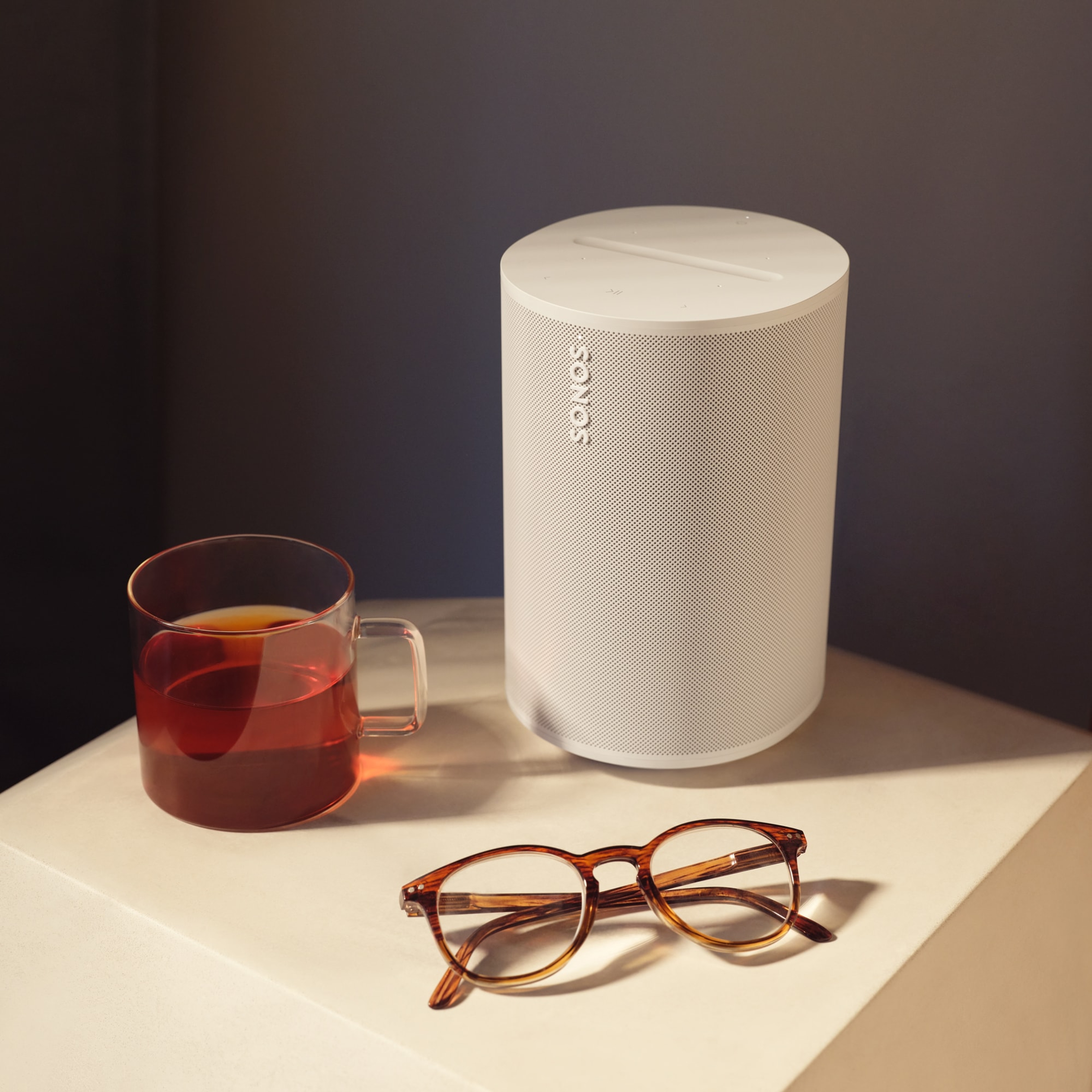 A white Era 100 speaker on a nightstand next to a cup of tea and reading glasses