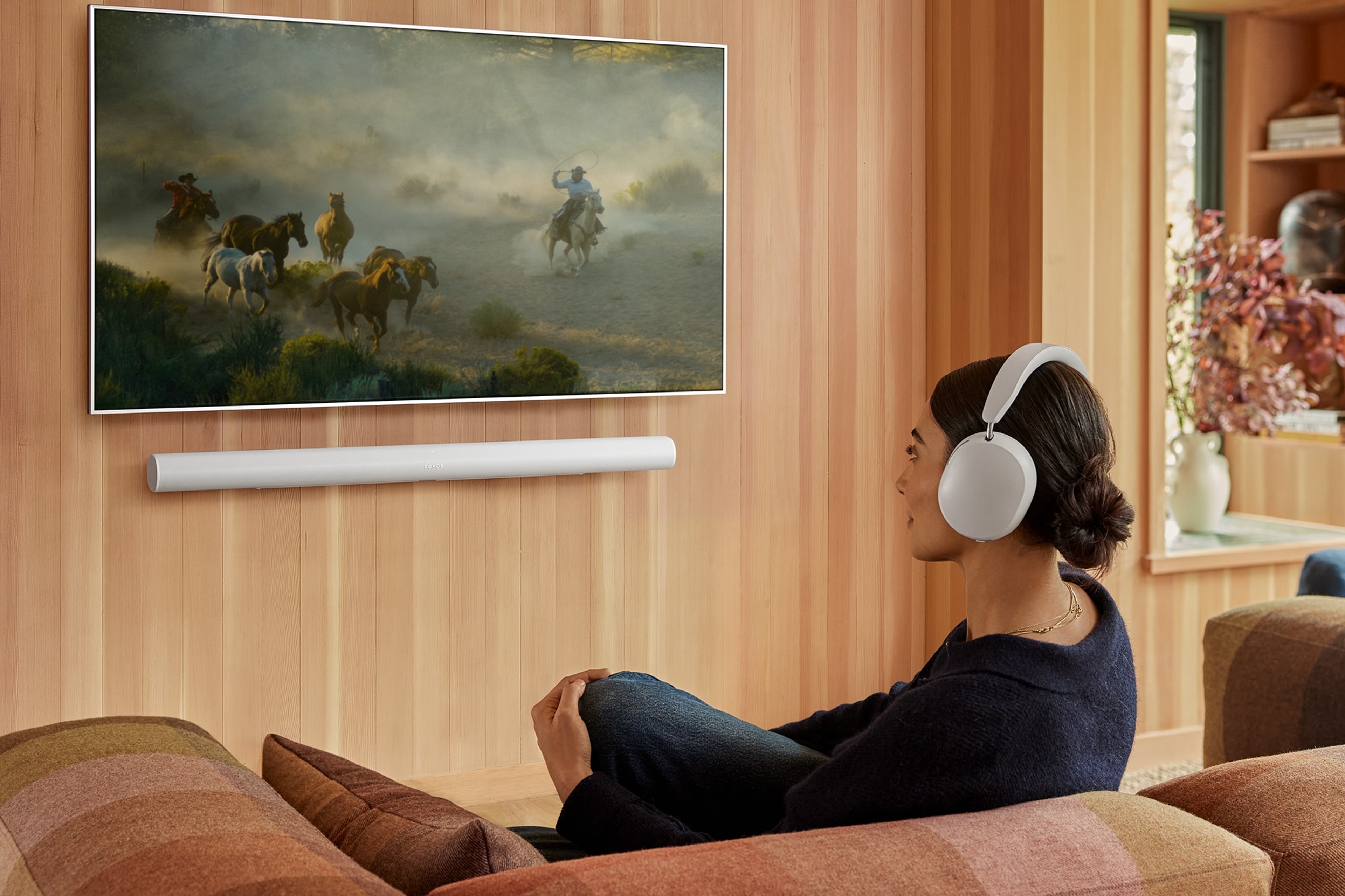 Person wearing white Sonos Ace headphones while watching TV with a white Arc Ultra mounted underneath