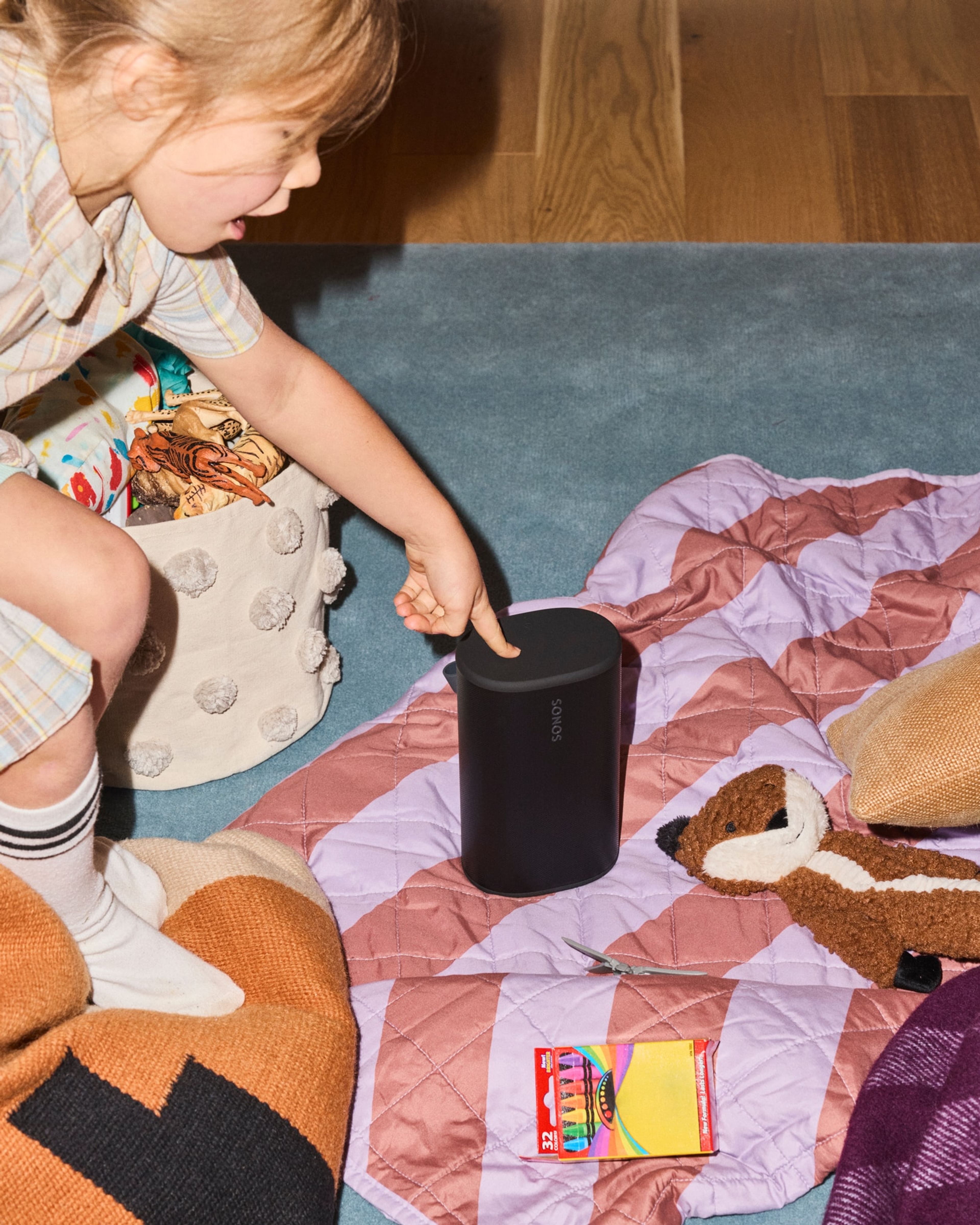 A young child on the floor with blankets and stuffed animals using the top controls on a black Sonos Play