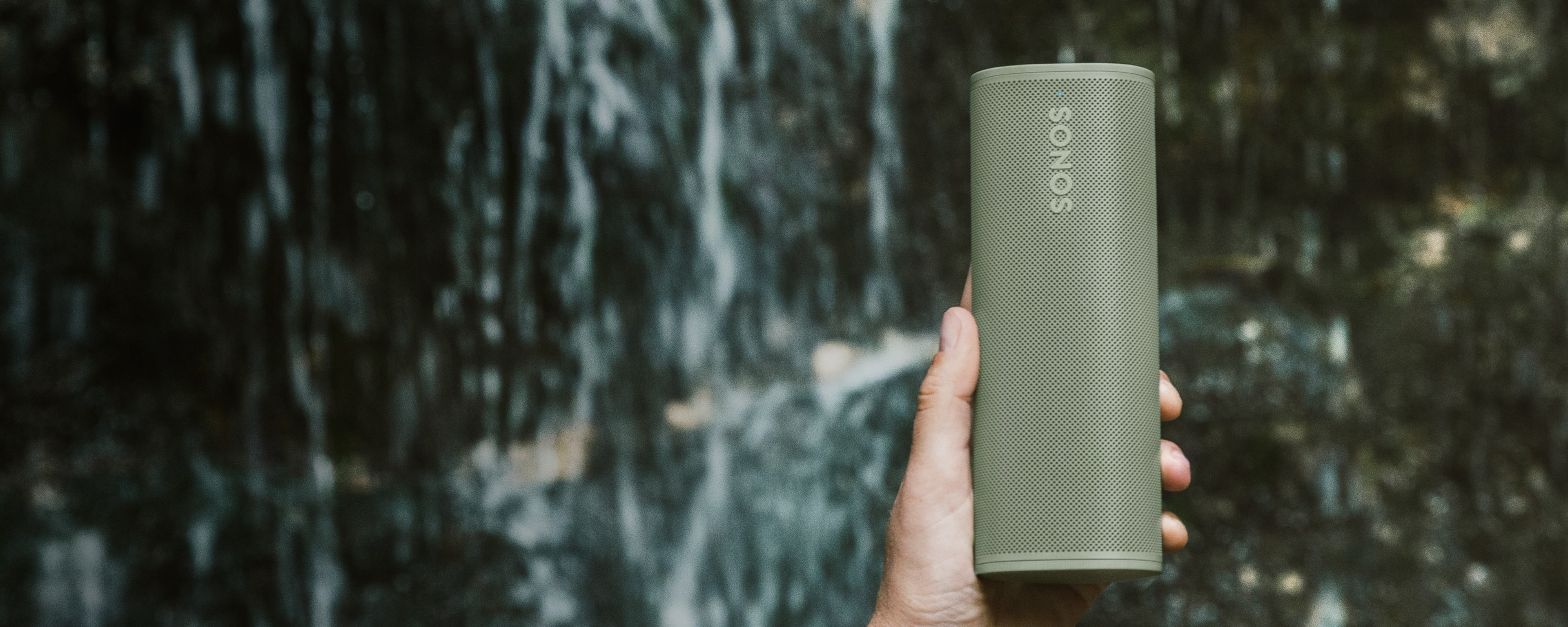 Una mano sostiene un Sonos Roam 2 en color Olive con una catarata al fondo