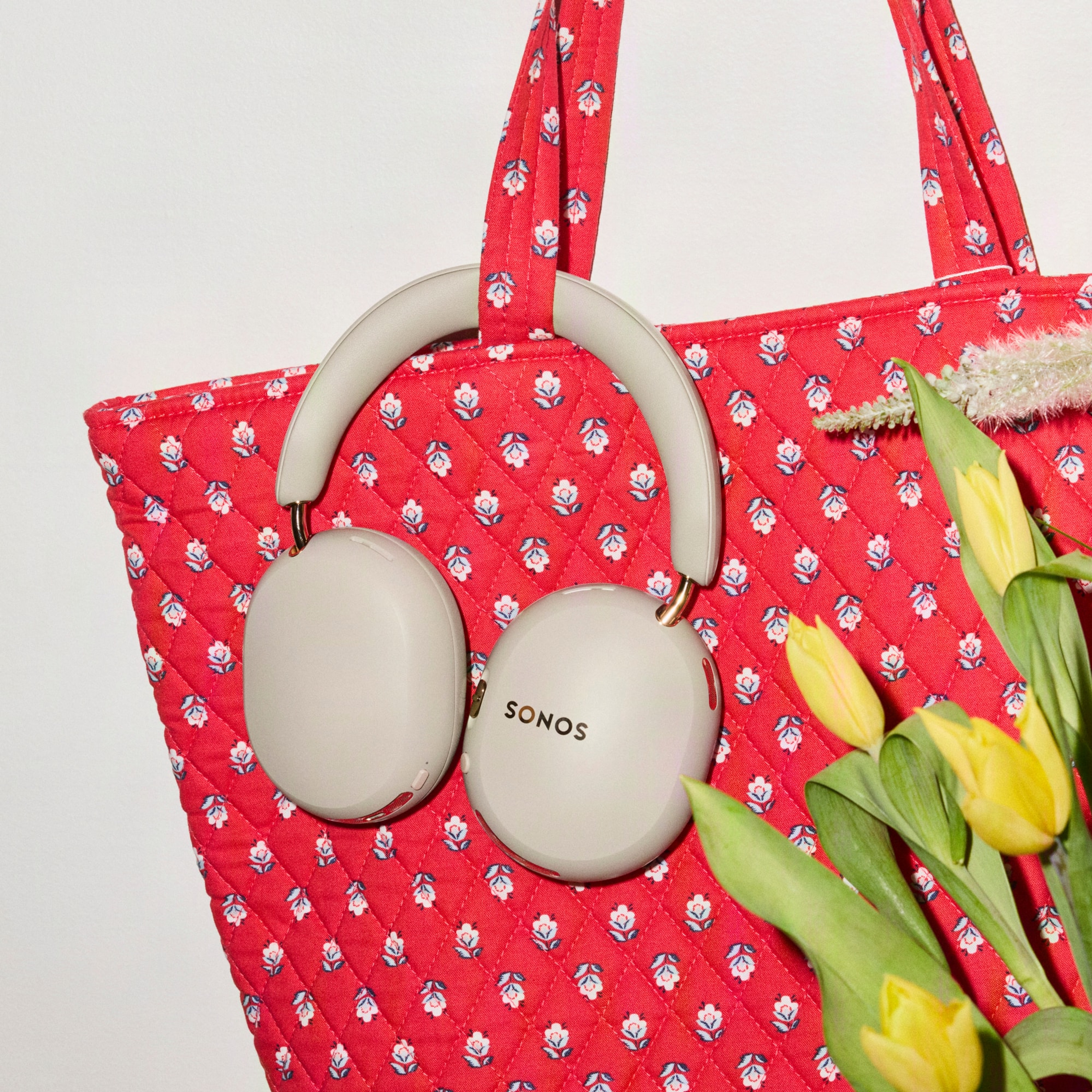 Sonos Ace Ultra headphones in sand, hanging on a red floral tote bag next to a vase full of yellow tulips