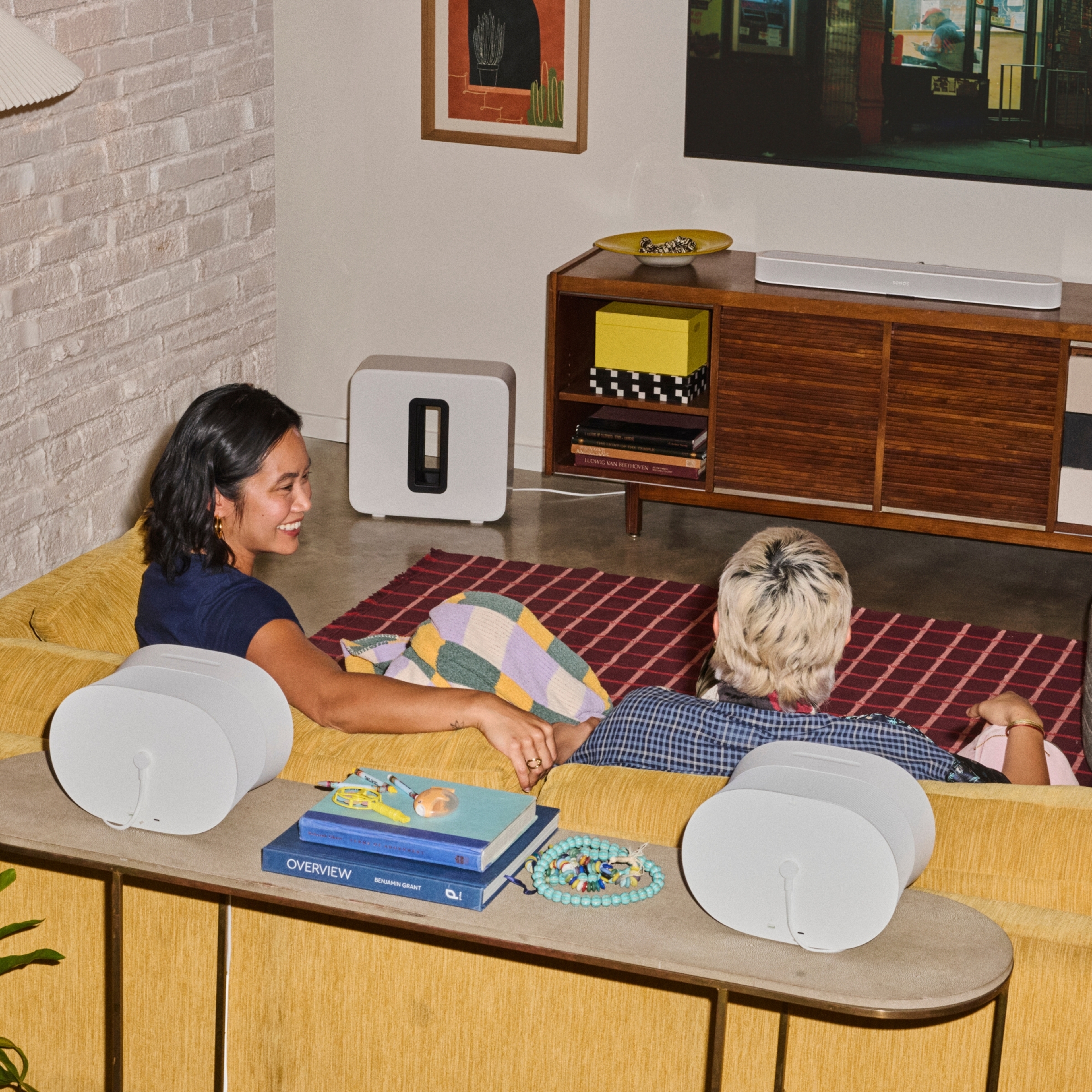 Two people sitting on a yellow couch with Era 300 speakers on a wooden console table in a living room with a white Sonos Beam Ultra and Sub 4 in the background